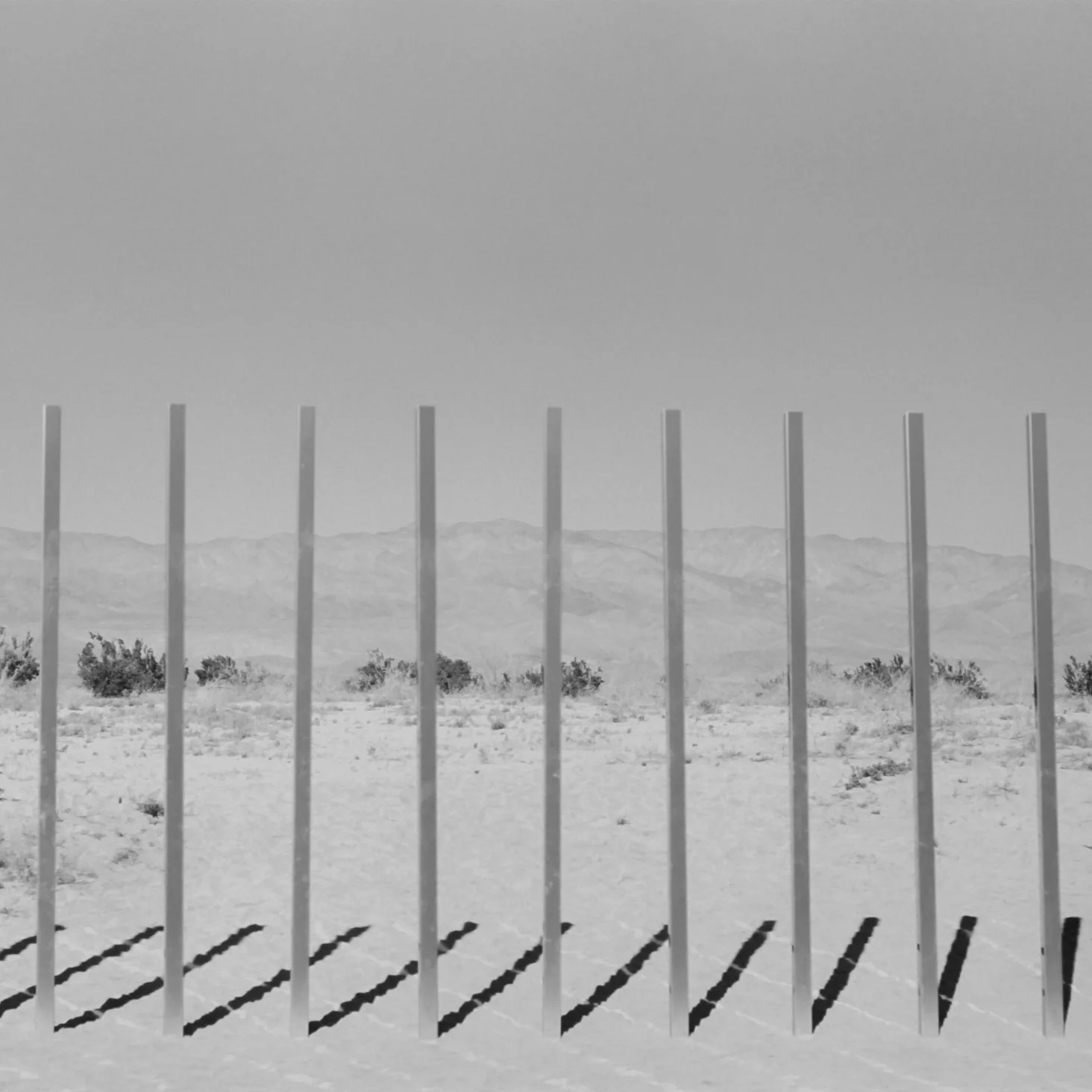 A black and white photo of a desert landscape with mountains in the distance, viewed through vertical metal fence posts casting shadows on the sandy ground.