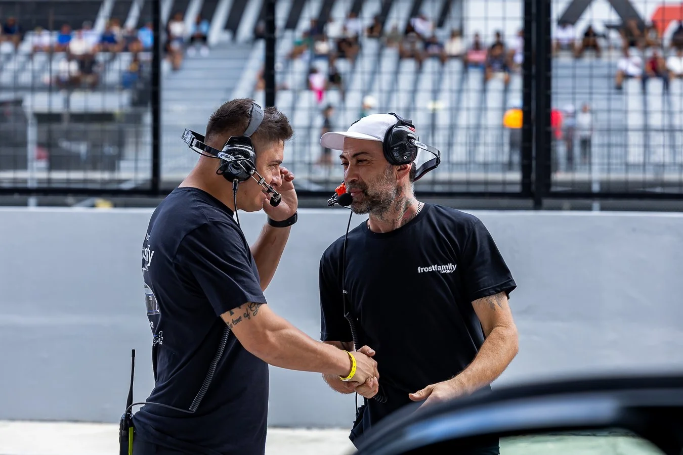 Two men at a racing event shaking hands, both wearing black shirts and headsets, with spectator stands in the background.