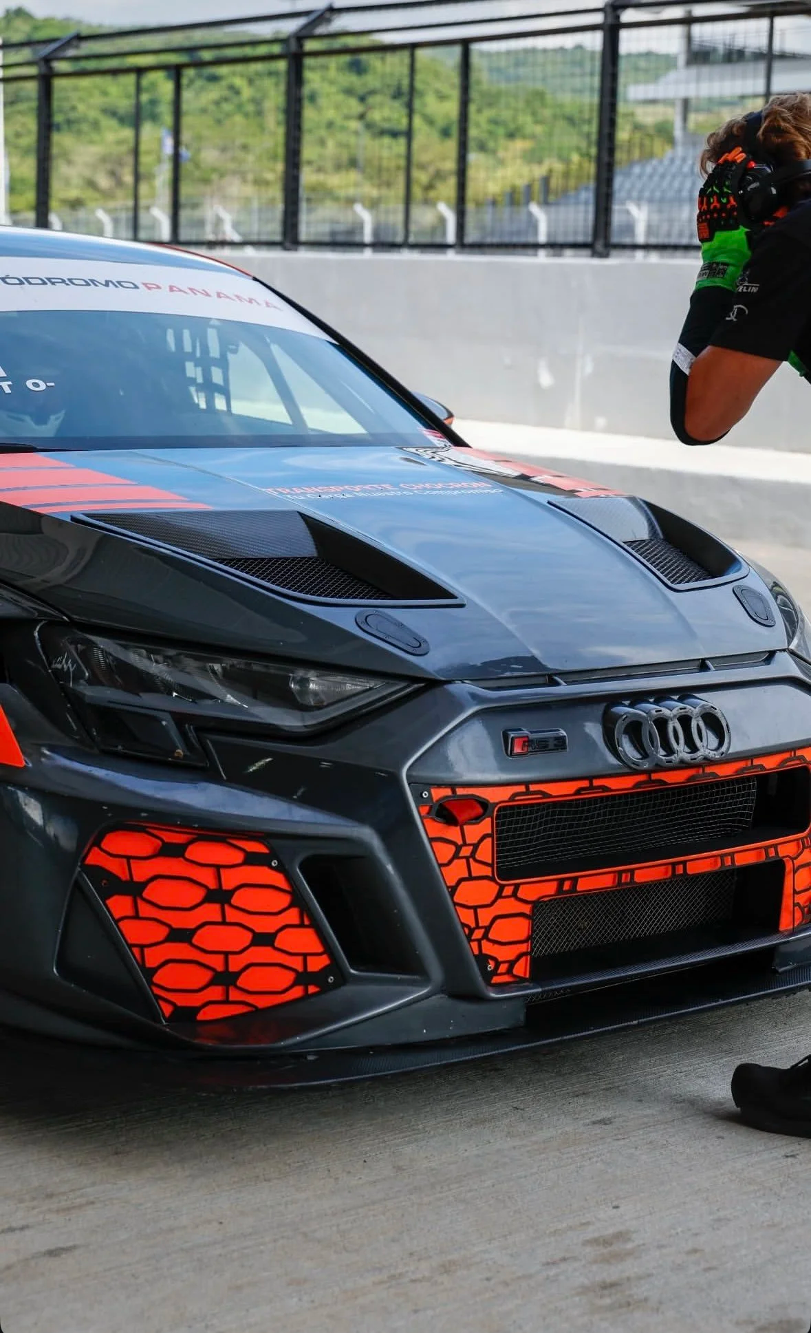 Close-up of an Audi racing car with black and red liveries, a custom grille, and a sleek aerodynamic design, parked in a racing pit with a crew member adjusting his helmet in the background.