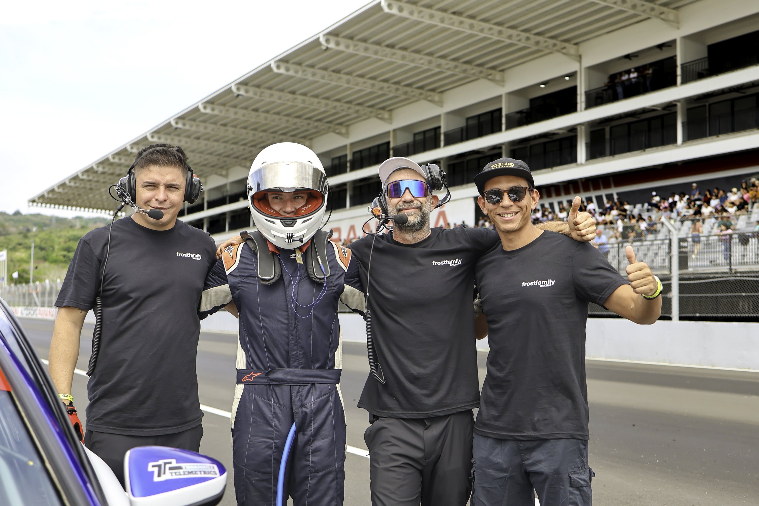 Four men standing together on a race track, posing for a photo, with a grandstand filled with spectators in the background. The second man is dressed in a racing suit and helmet, while the other three are wearing matching black shirts with "frostfamily" written on them. Two of them are giving thumbs-up signs and all are smiling.