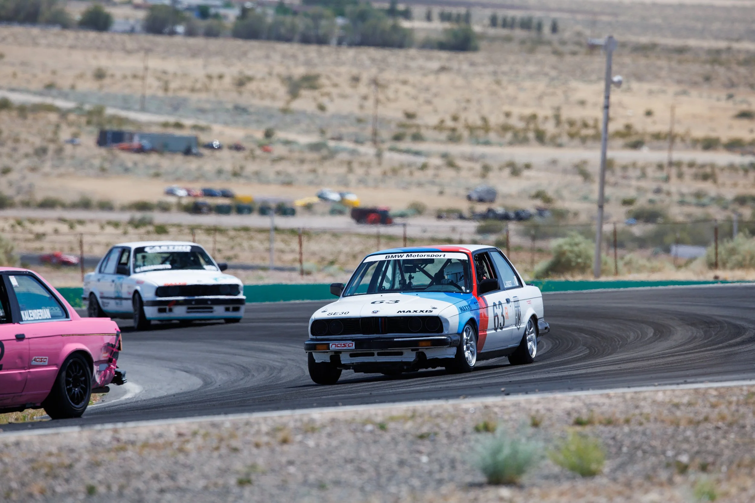 Race cars on a dirt track in desert landscape with brown hills in the background, including a BMW Motorsport car, a pink car, and a white car.