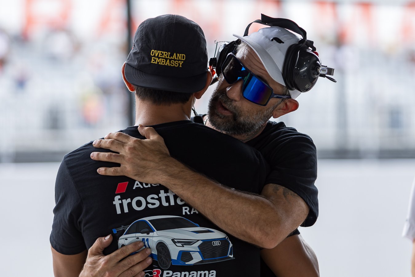 Two men hugging at a racing event. One man wears a cap with 'Overland Embassy' and a shirt with a racing logo. The other man wears racing goggles, a cap, and headphones.