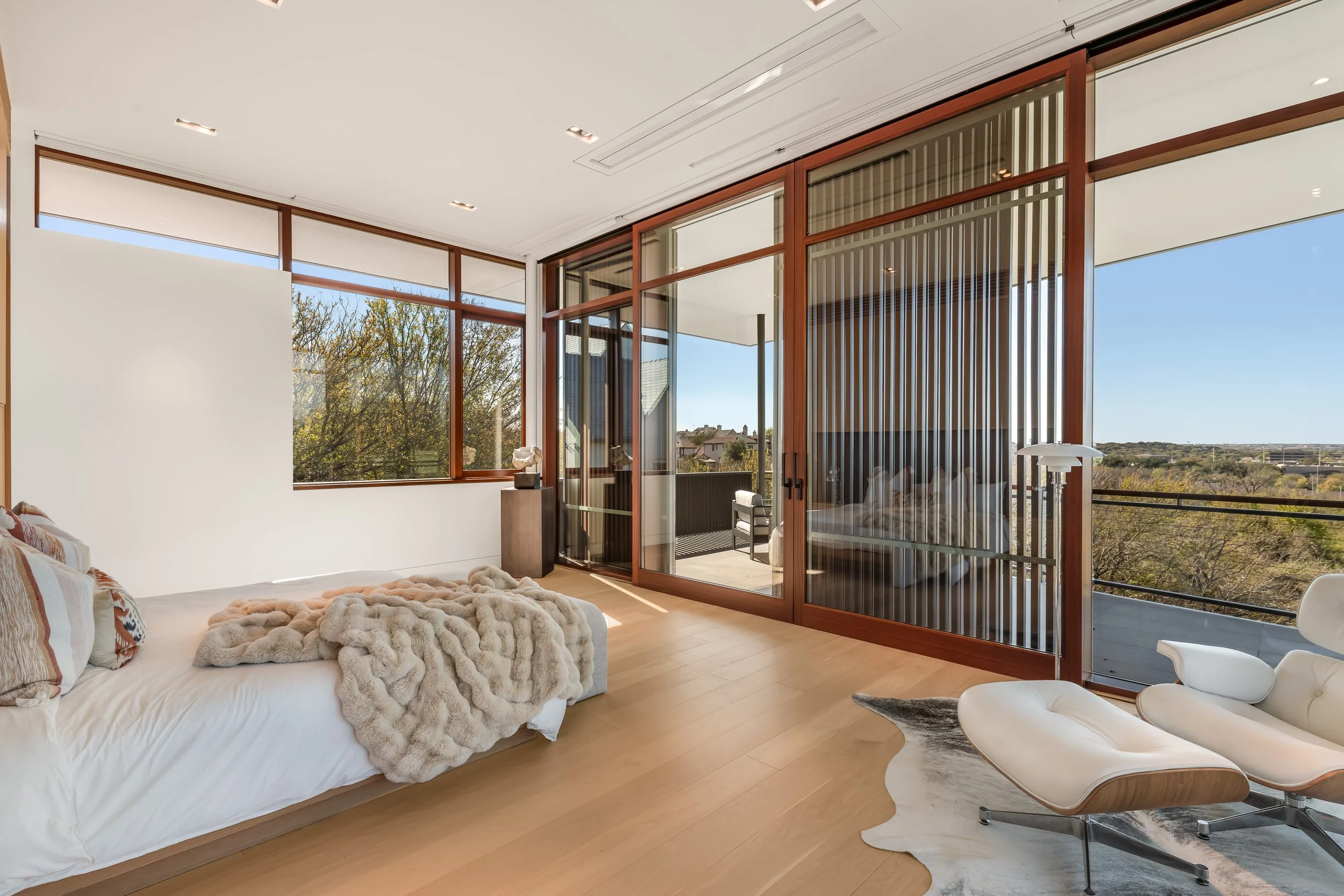 Modern bedroom with large windows, a balcony, a white bed with fur throw, and an Eames lounge chair.
