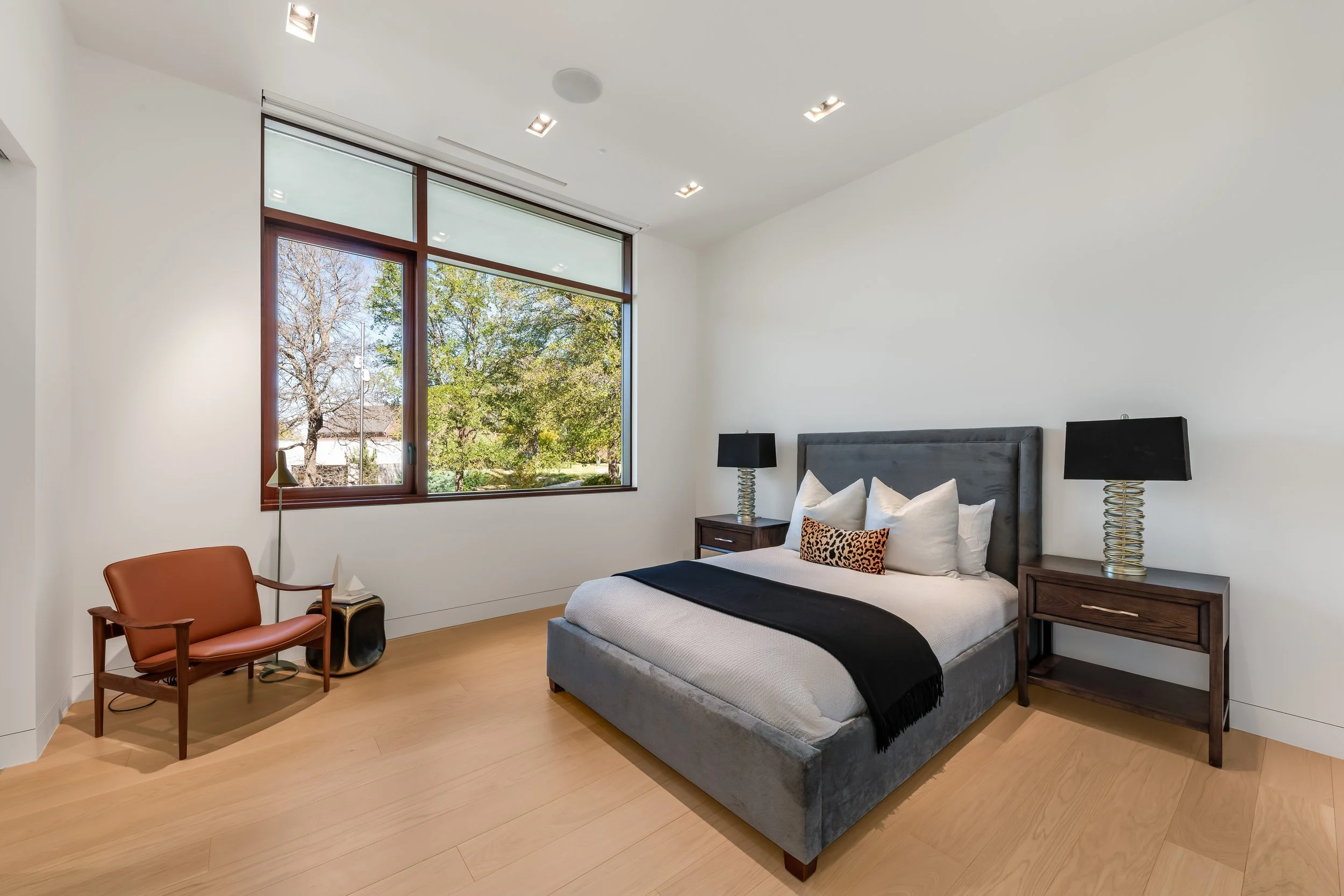A modern bedroom with a large window showing trees outside, a queen-sized gray bed with white pillows and black blanket, two wooden nightstands with black lamps, a wooden chair, and a small side table.