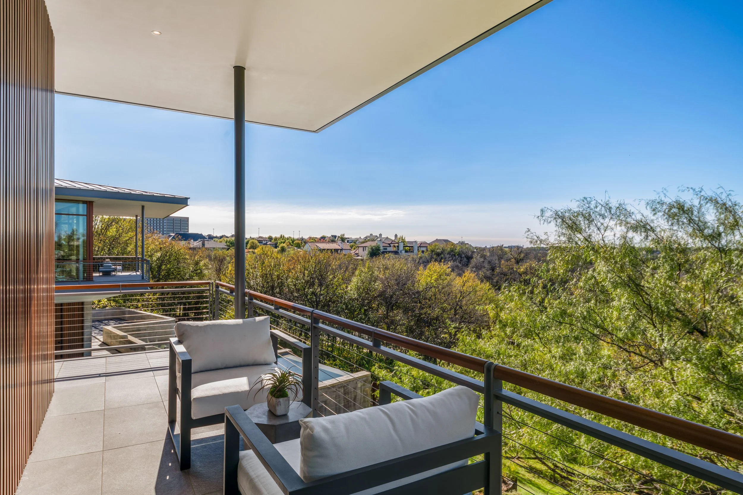 Balcony with two outdoor chairs and a small table holding a potted plant, overlooking a cityscape with trees and buildings under a clear blue sky.
