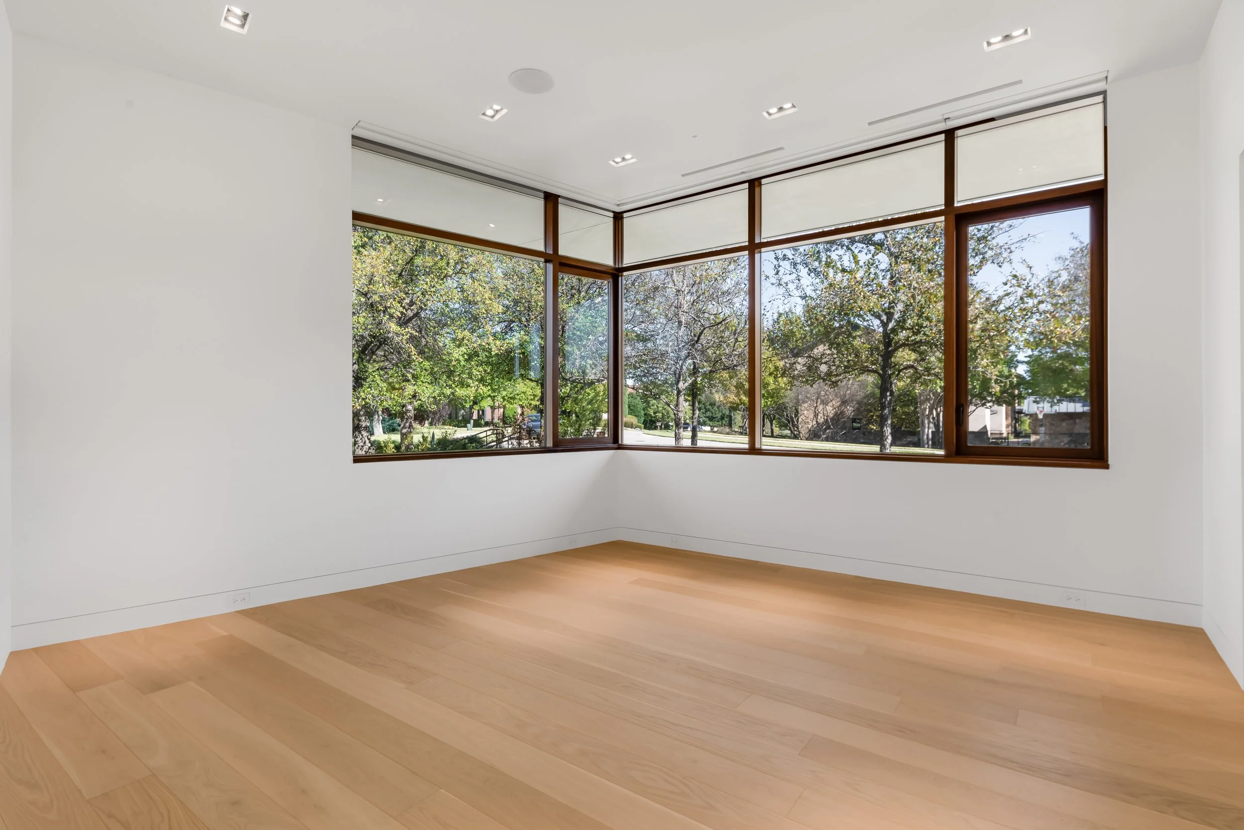 Empty room with large windows showing trees and greenery outside, white walls, wood flooring, and ceiling lights.