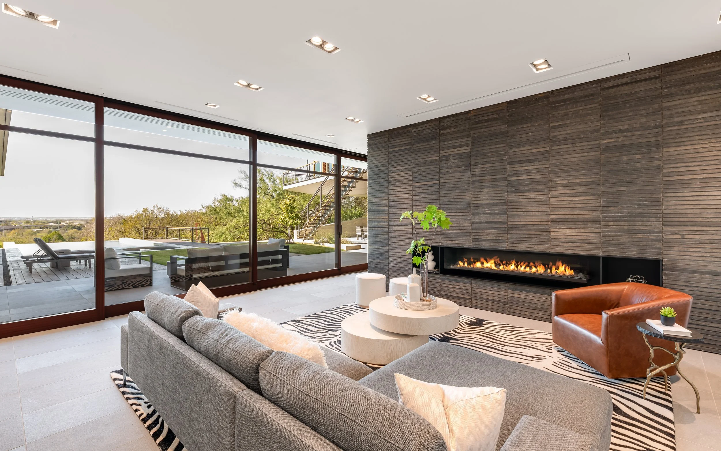 Modern living room with large windows, gray sectional sofa, orange armchair, fireplace, and zebra print rug.