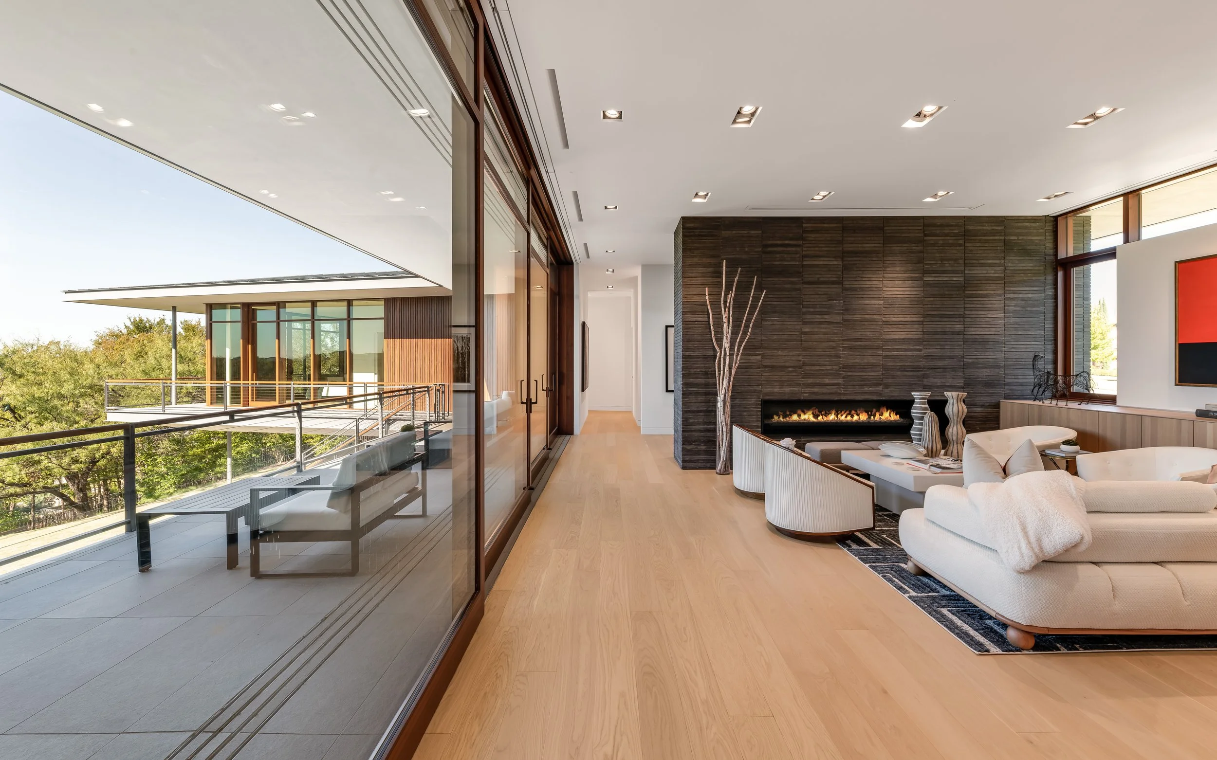 Modern living room with wooden flooring, a white sectional sofa, and a black accent wall with a linear fireplace. Large windows and glass doors allow natural light, with an outdoor balcony visible.