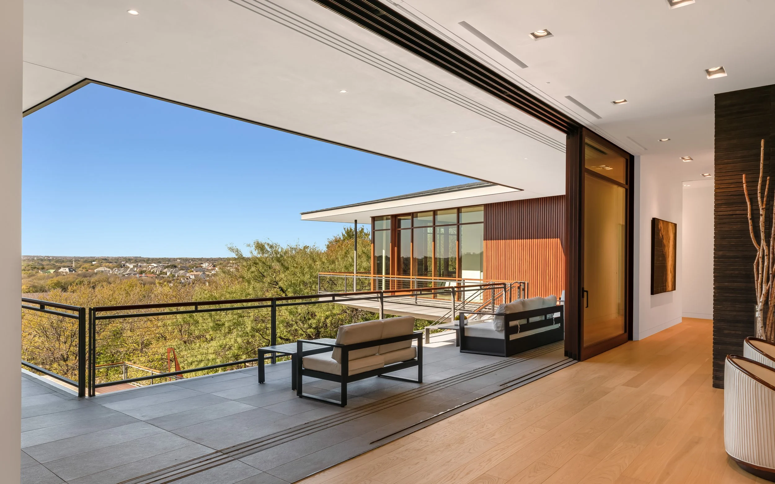 Interior view of a modern living space with an open sliding glass door leading to a balcony. The balcony has outdoor seating and offers a view of trees and a distant neighborhood under a clear blue sky.