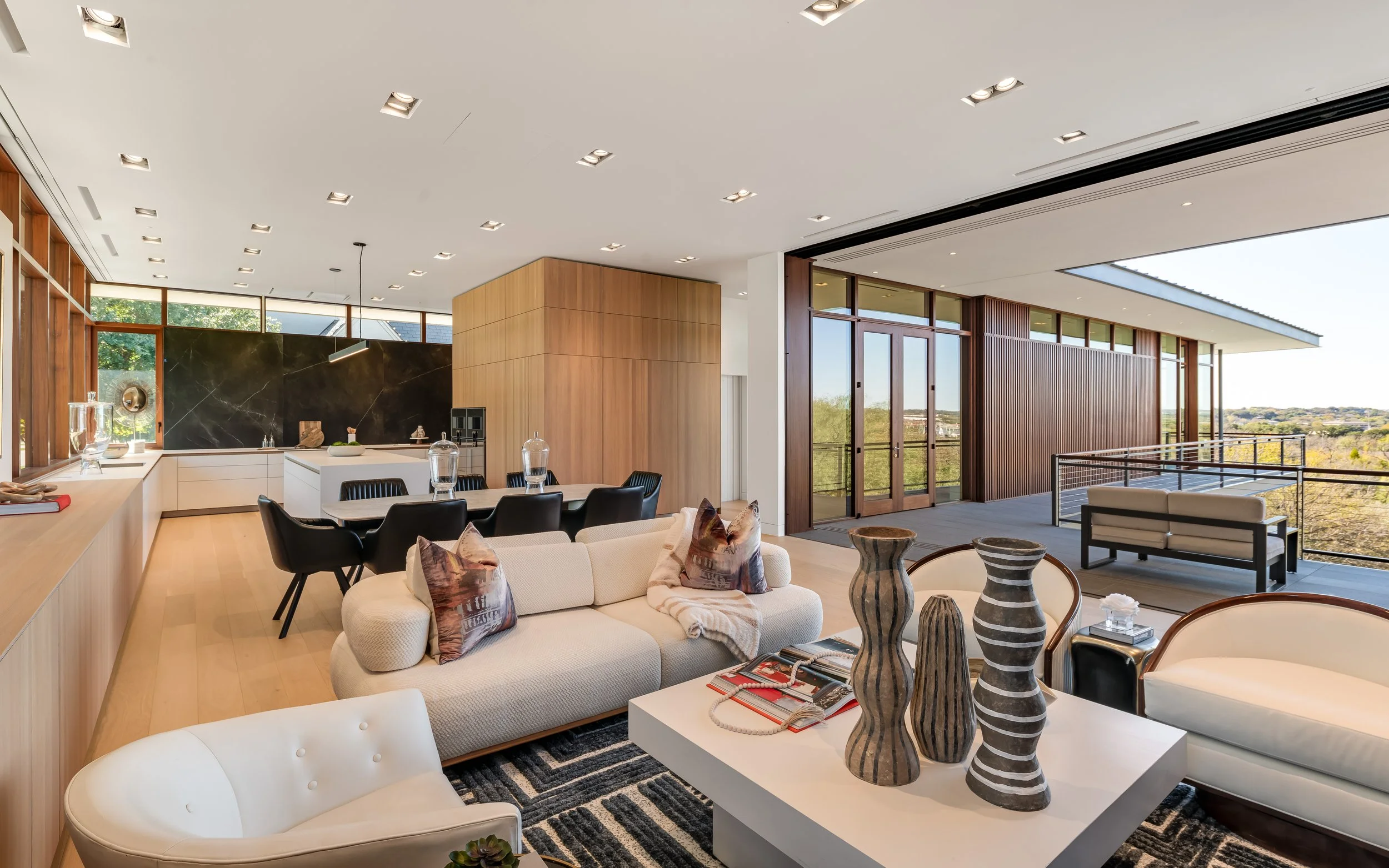 Modern living room with open floor plan, large windows, and minimalist decor. Includes seating area with sofa and chairs, a kitchen with island, and a spacious outdoor balcony.