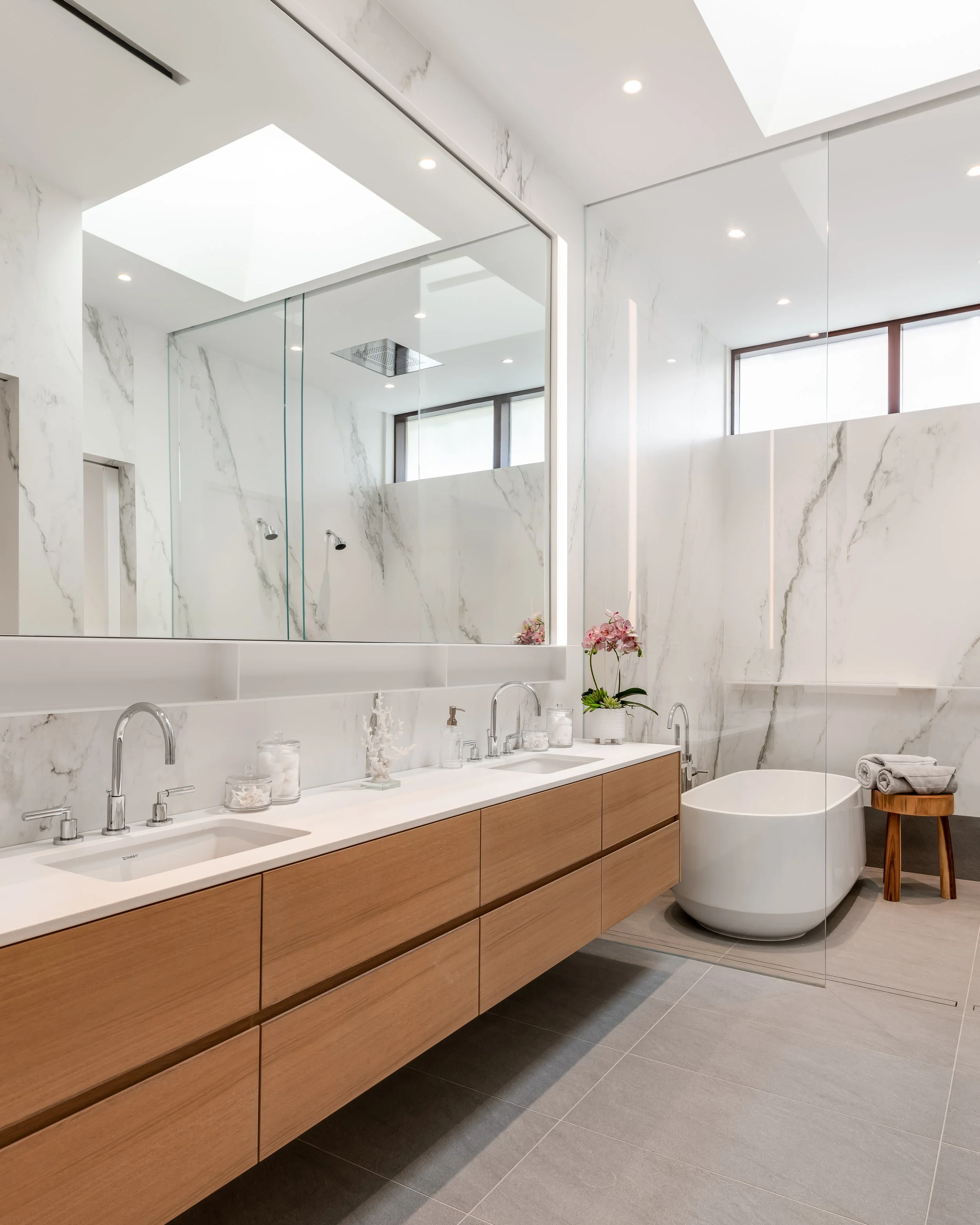 Modern bathroom with wooden vanity, double sinks, large mirror, bathtub, pink orchids, gray floor tiles, marble walls, and skylight