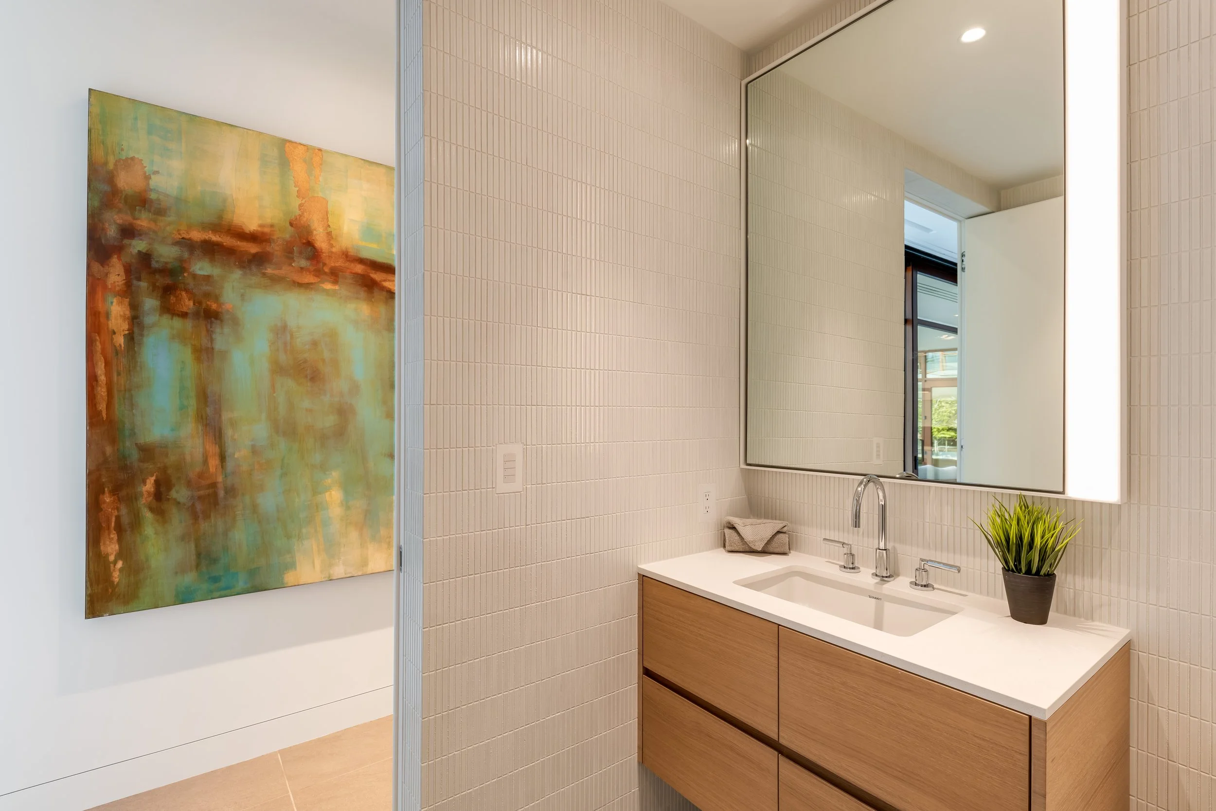 Modern bathroom vanity with a large mirror, white countertop, and two drawers, decorated with a small potted plant and a folded towel, adjacent to an abstract painting on a white wall.