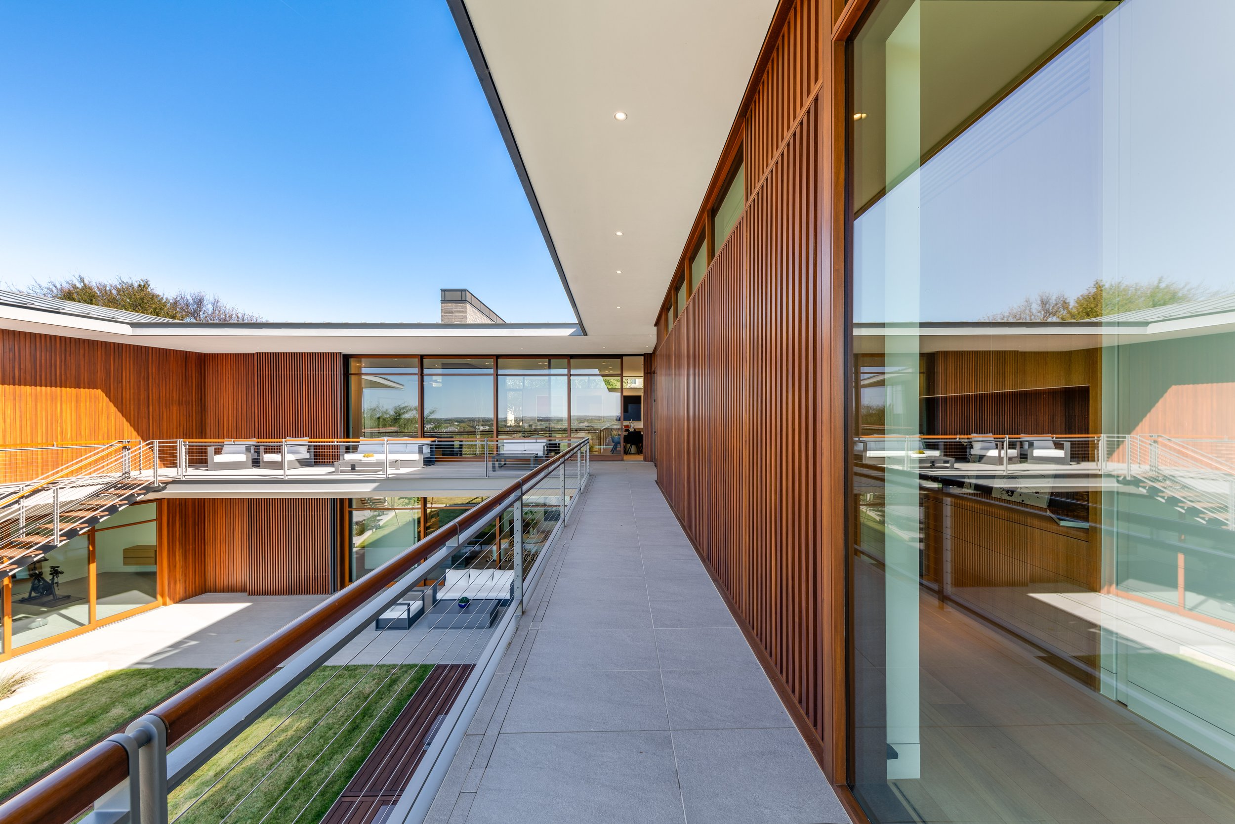 Modern building with glass walls, wooden vertical slats, outdoor balcony, and seating area, under a clear blue sky.