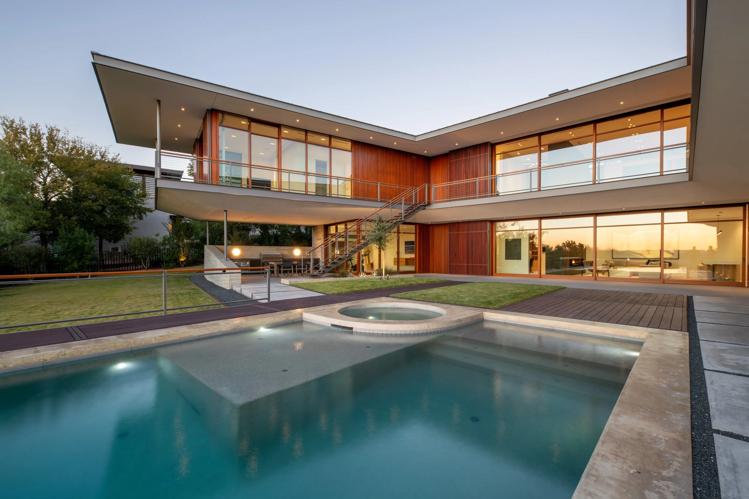 Modern two-story house with large glass windows, outdoor pool, hot tub, and patio area during sunset