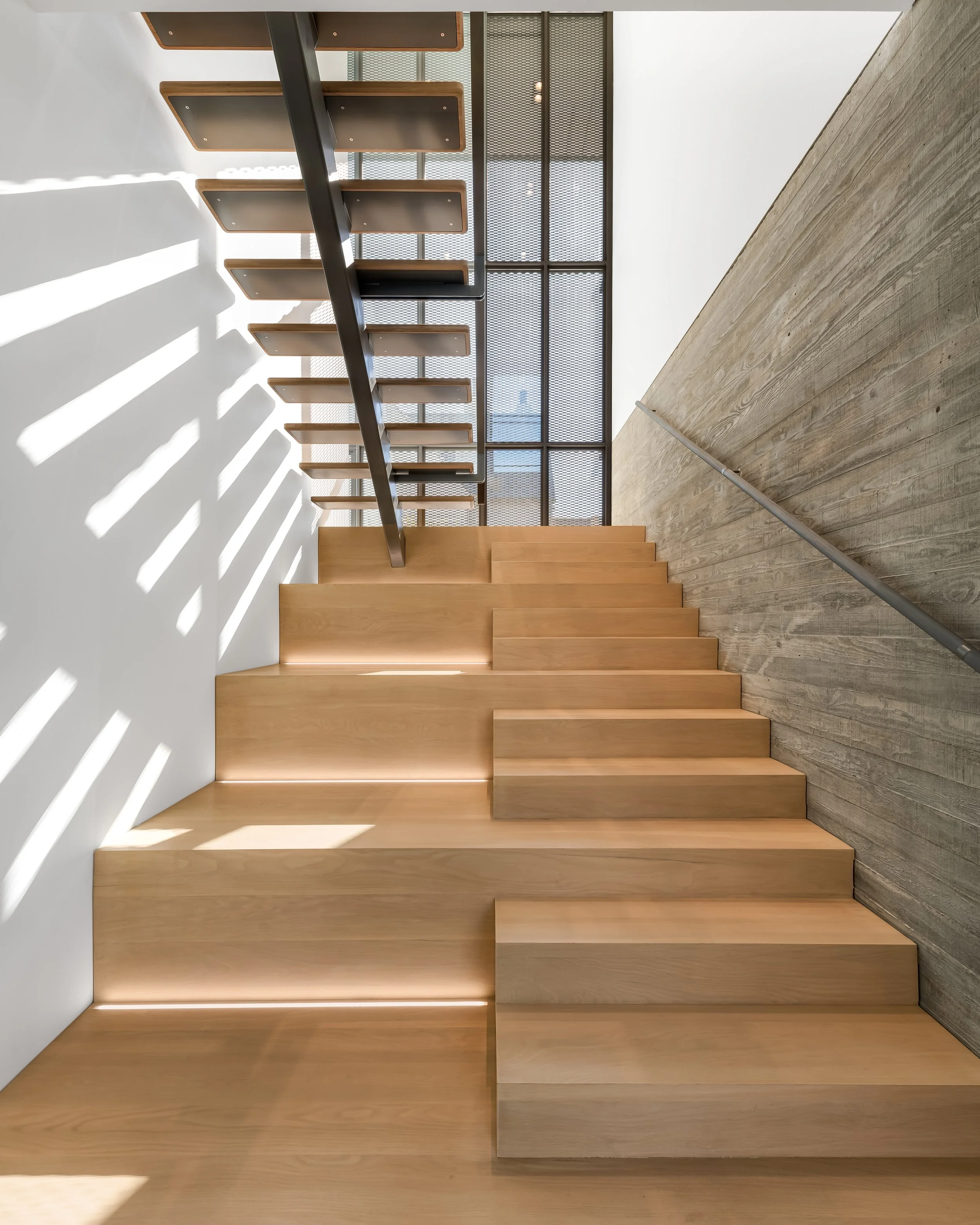 Modern staircase with wooden steps, metal handrail, and natural light casting striped shadows on the wall.