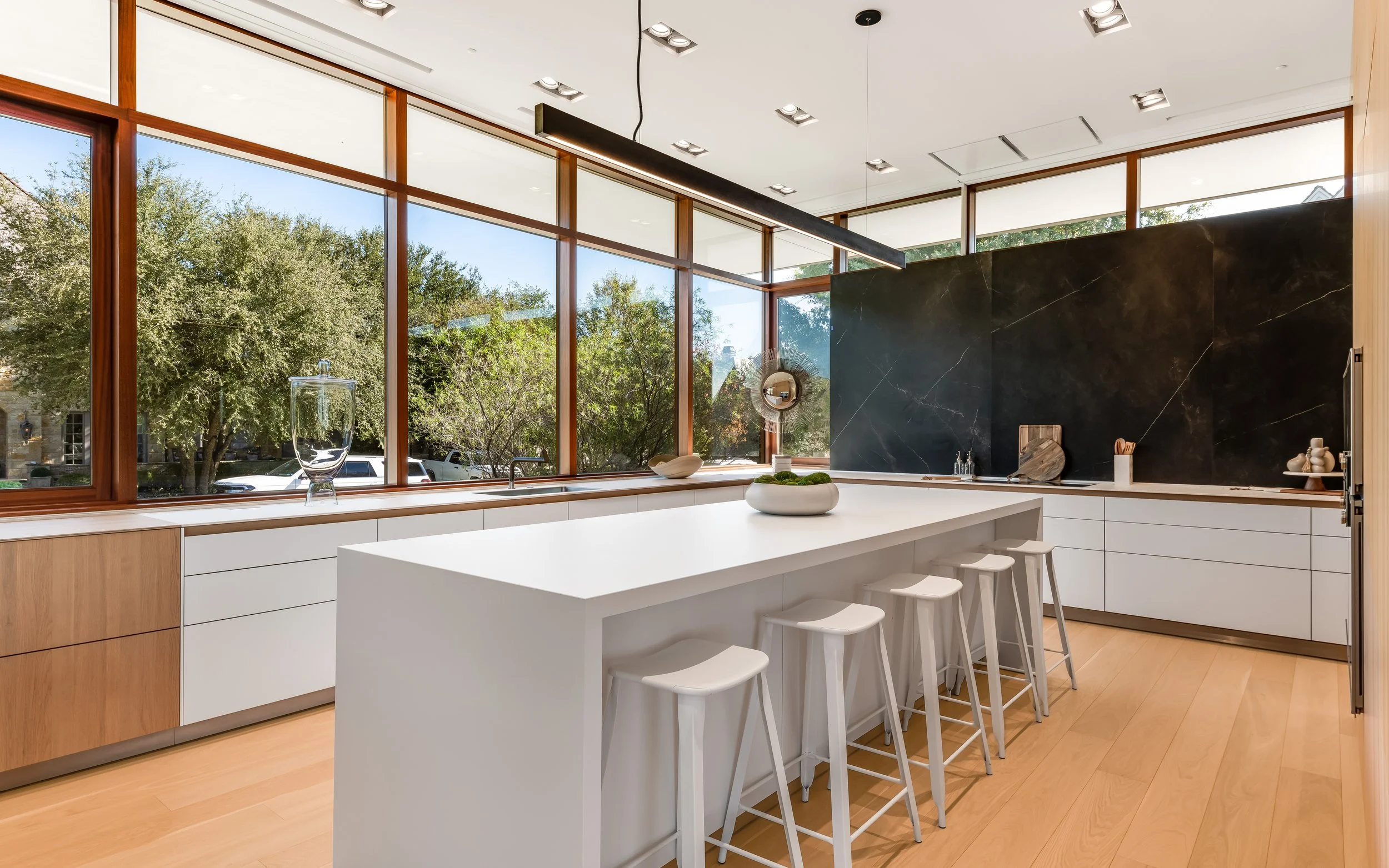 Modern kitchen with large windows, white island counter with six bar stools, black marble wall, light wood floors, and minimal decor.