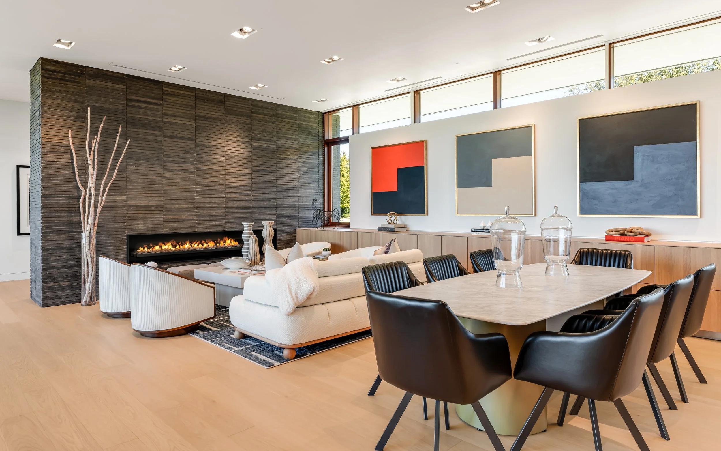 Modern living room with a white L-shaped sofa, black dining chairs around a marble table, a wall-mounted fireplace, and two large abstract paintings on the white wall.