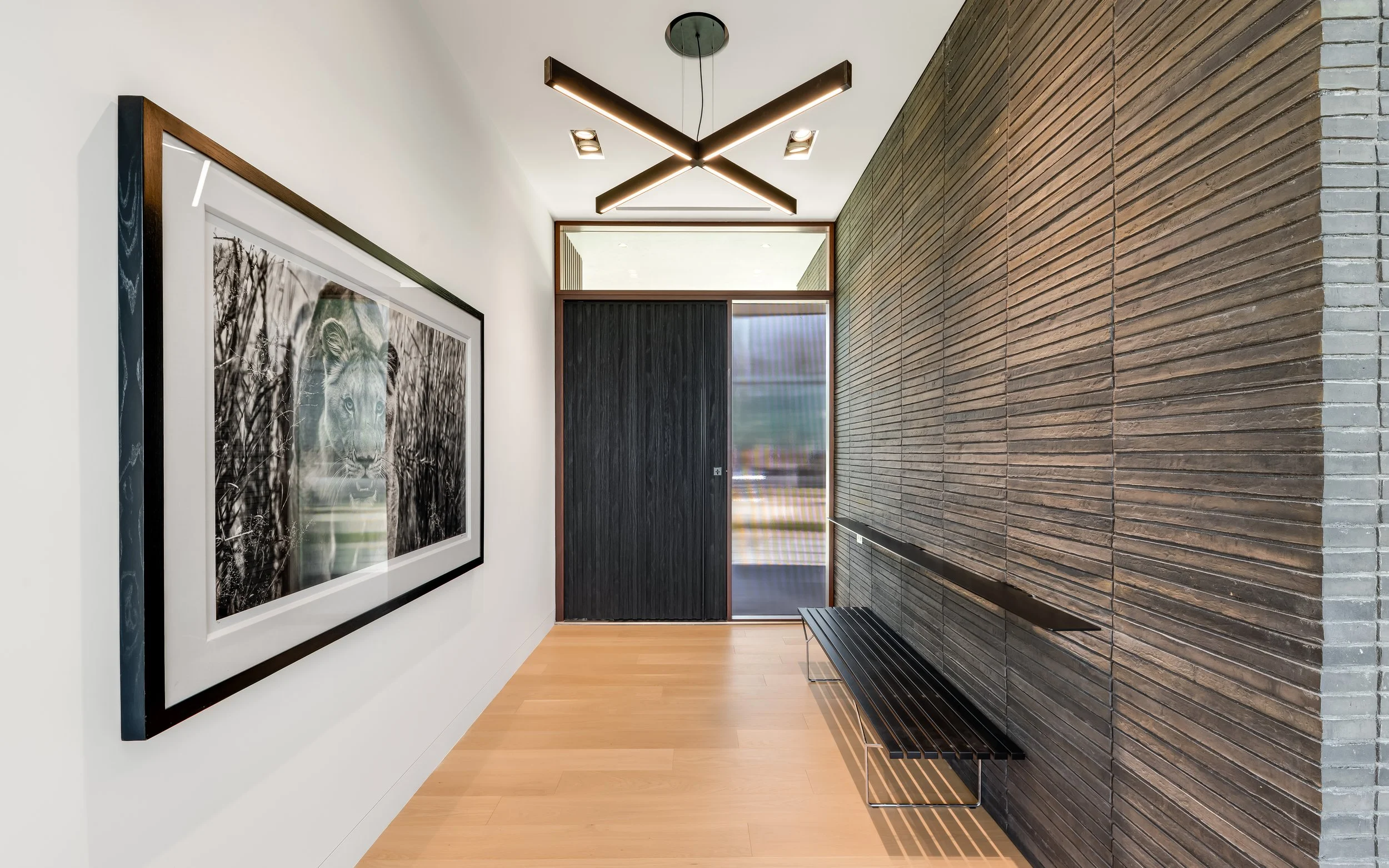 Modern hallway with a black and white lion photograph on the wall, wooden floor, a black bench, textured gray brick wall, and geometric ceiling lights.