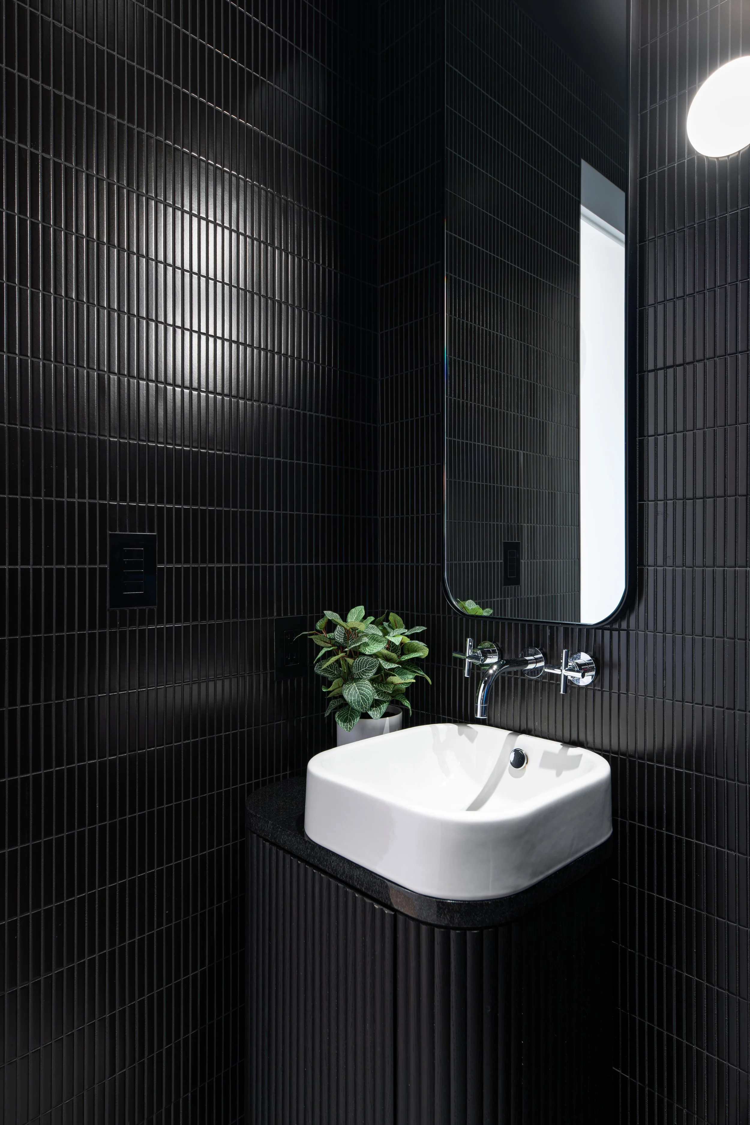 A modern bathroom with black tiled walls, a white vessel sink, a large mirror, and a small potted plant beside the sink.