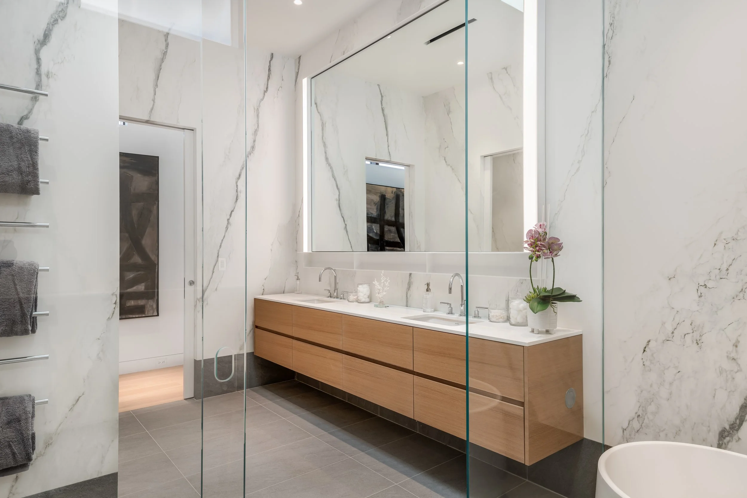 Modern bathroom with a double sink vanity, large mirror, marble walls, gray floor tiles, and a potted orchid flower.
