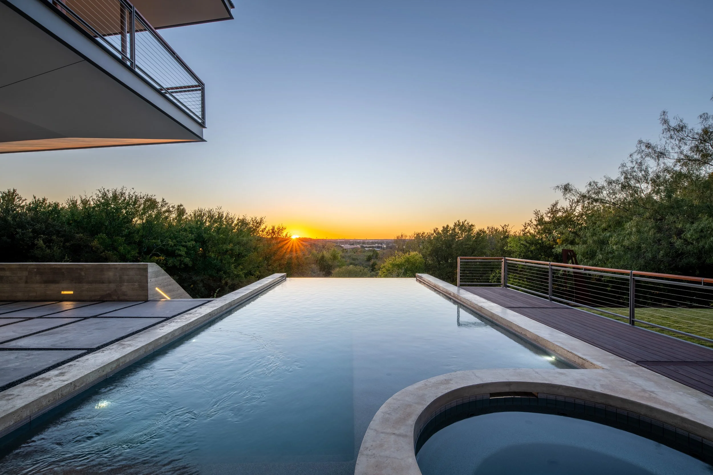 Luxury modern outdoor pool and hot tub area with a view of the sunset over trees, adjacent to a balcony with metal railing and wooden deck.