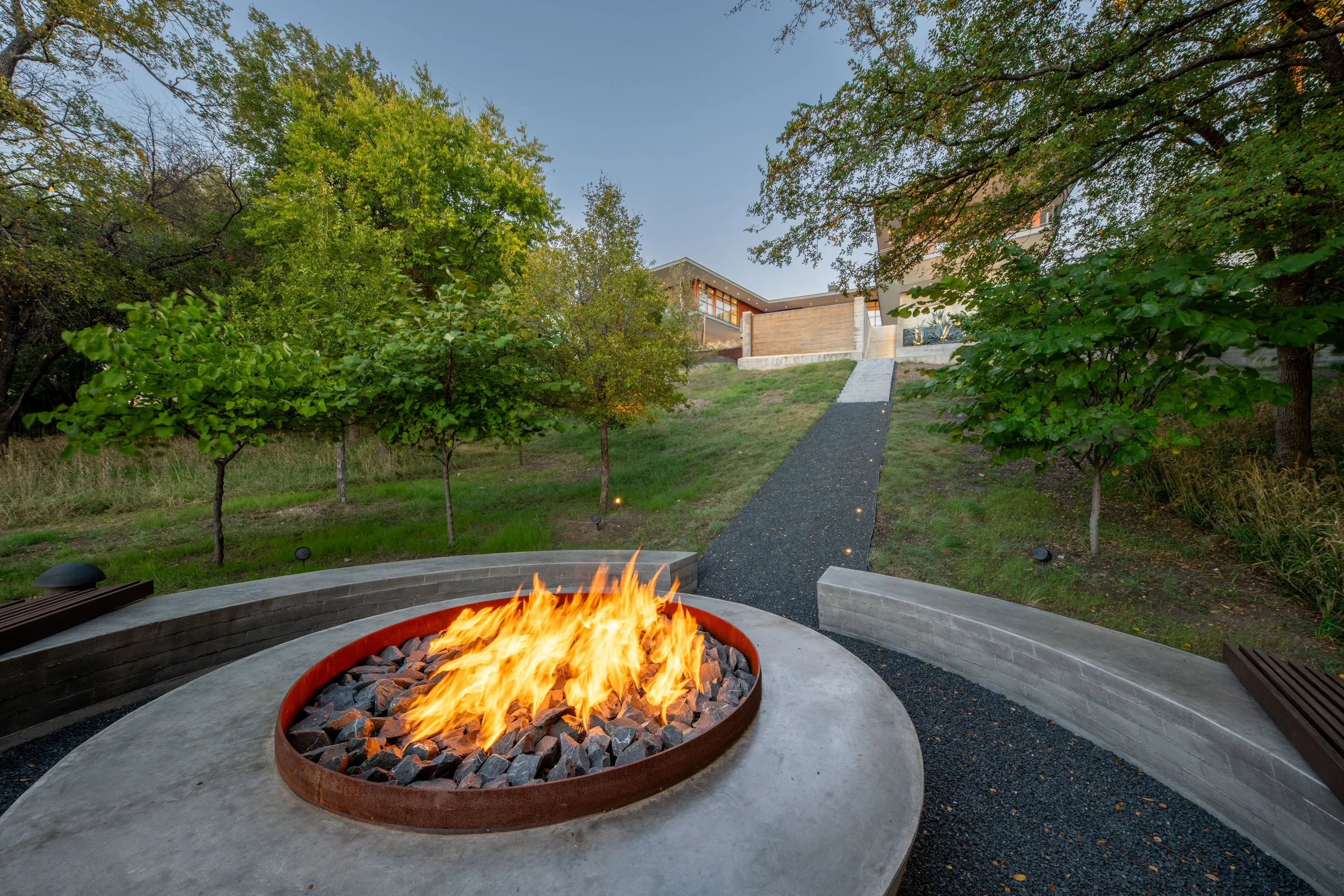 View of a modern backyard with a fire pit and a pathway leading up a hill towards a house, surrounded by greenery and trees.