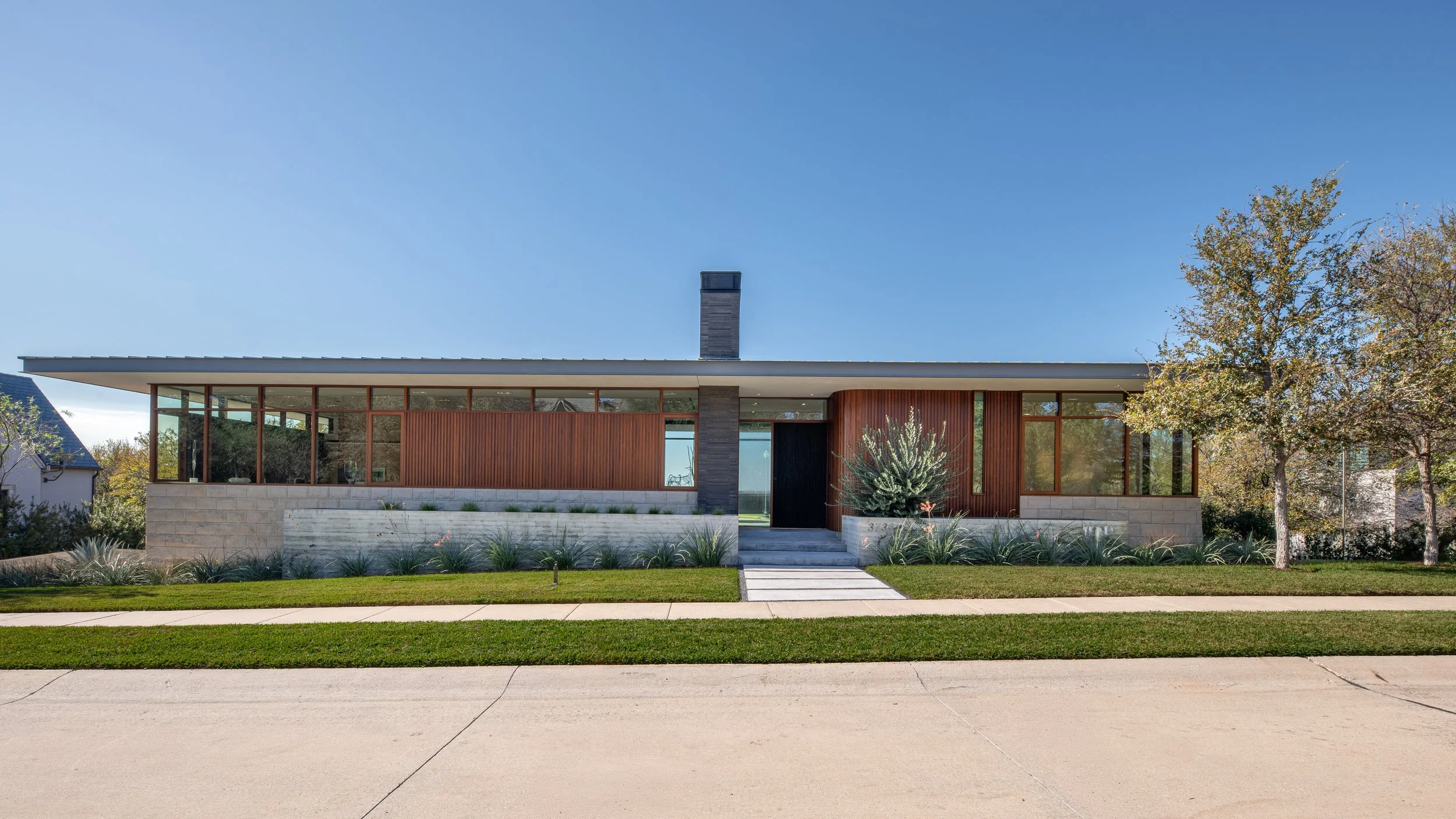 Modern single-story house with large glass windows, wooden paneling, and a central chimney, surrounded by a landscaped lawn, trees, and a sidewalk under a clear blue sky.