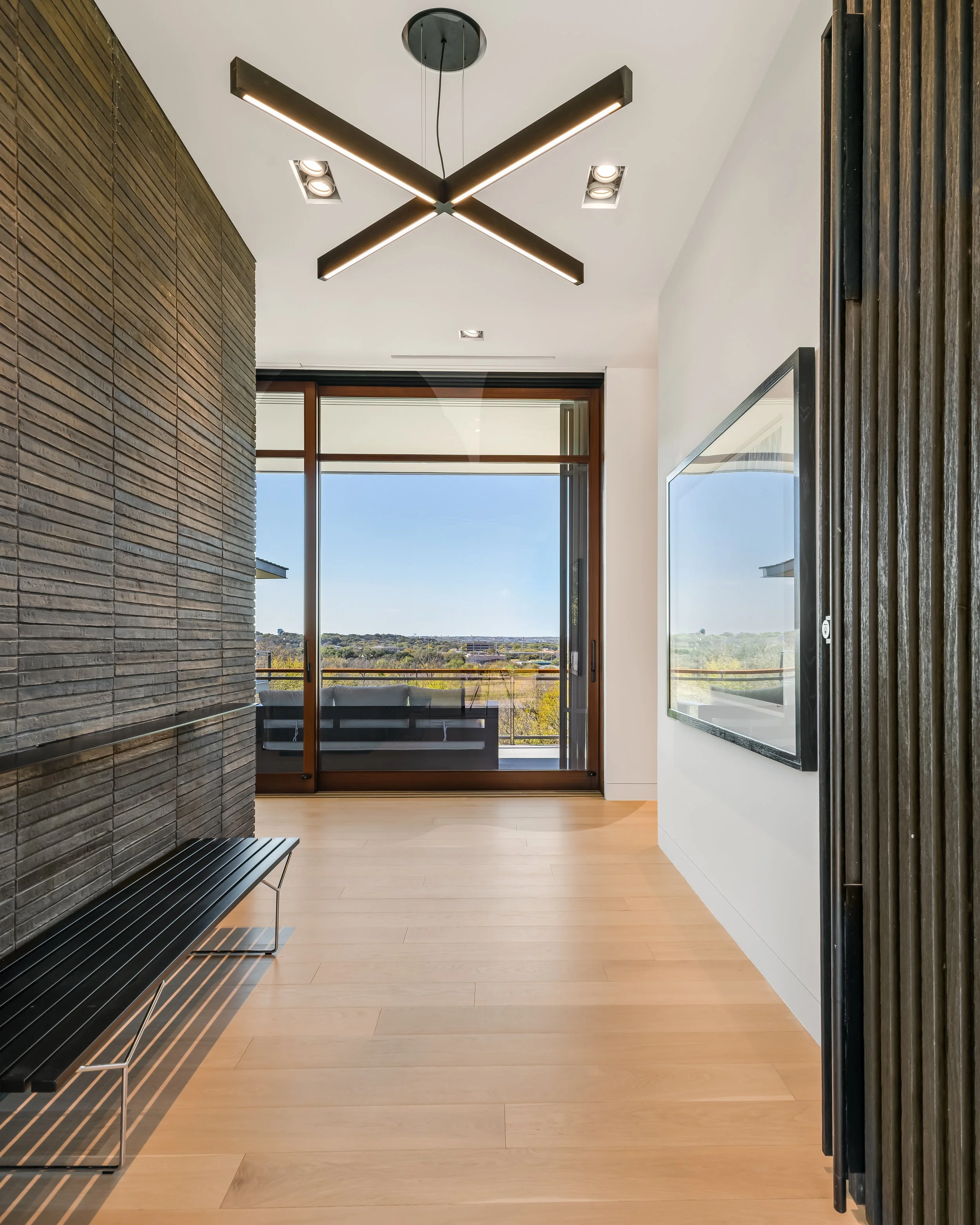 Modern living room with large glass sliding door leading to balcony, view of landscape outside, black bench, wood flooring, textured black wall, flat-screen television on white wall, unique black ceiling light fixture with angled bars.