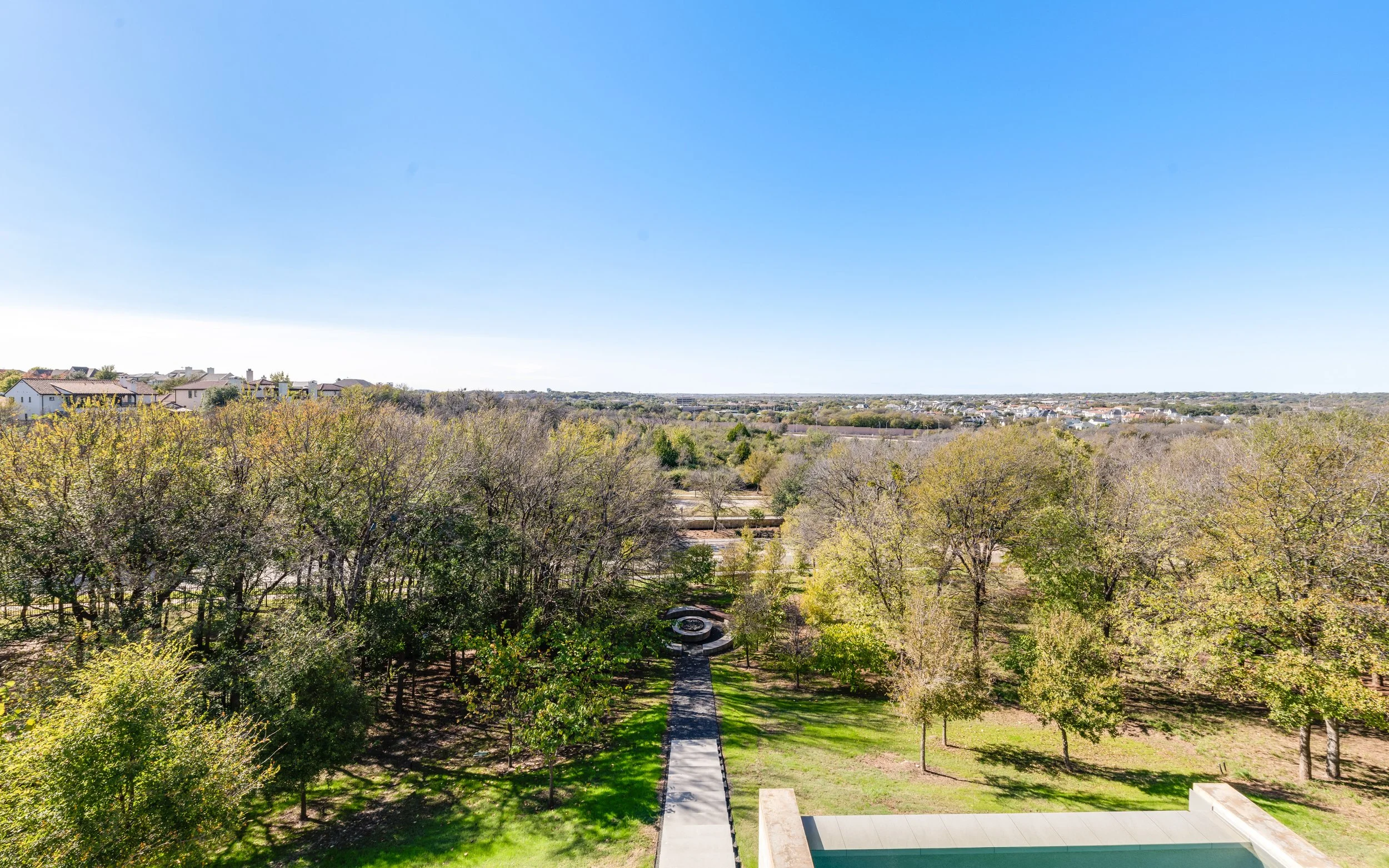 Aerial view of a park with a walking path, surrounding trees, and a fountain, with houses and a cityscape in the distance under a clear blue sky.