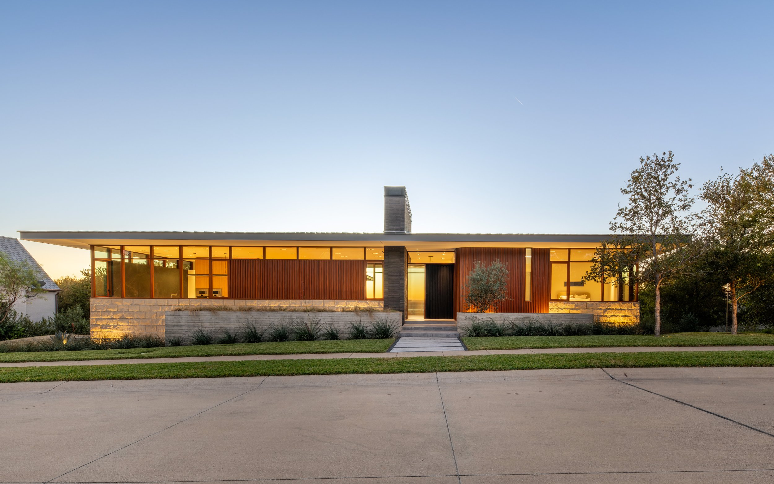 Modern single-story house with large glass windows, wood paneling, and a stone foundation, set against a clear evening sky.