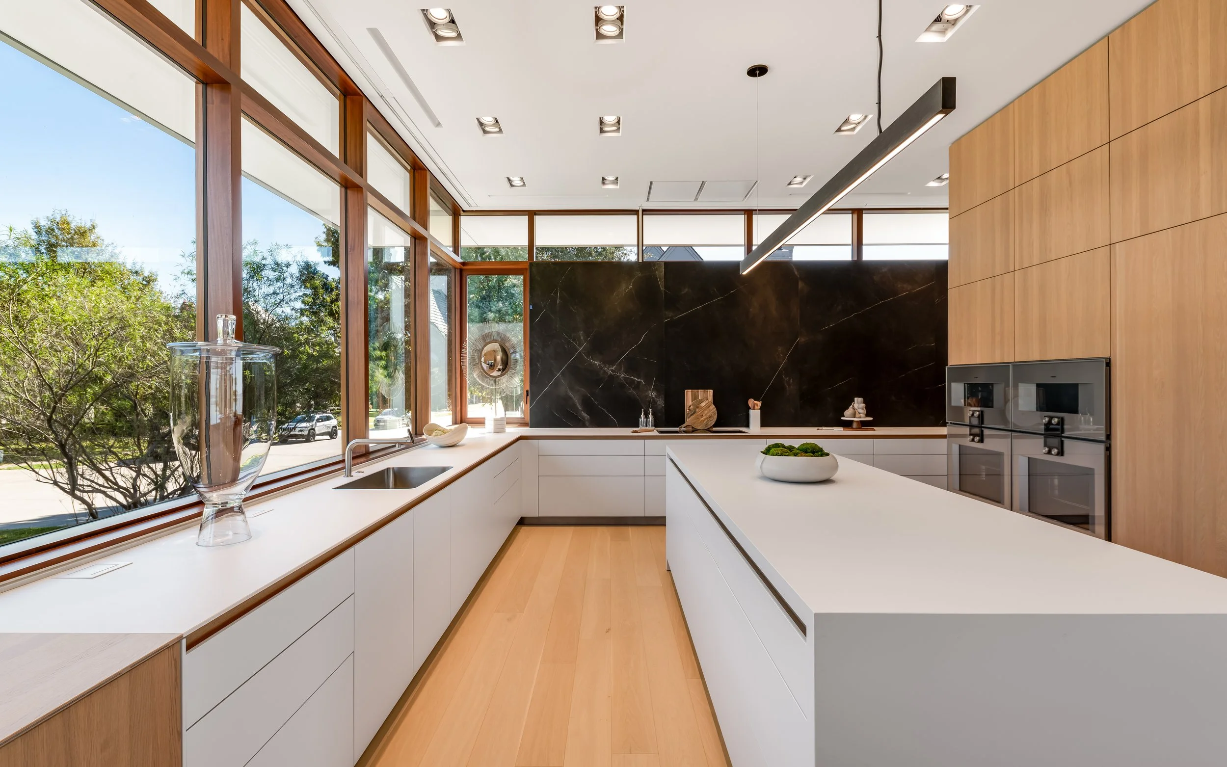 Modern kitchen with large windows, white countertops, wood floors, and black marble backsplash.