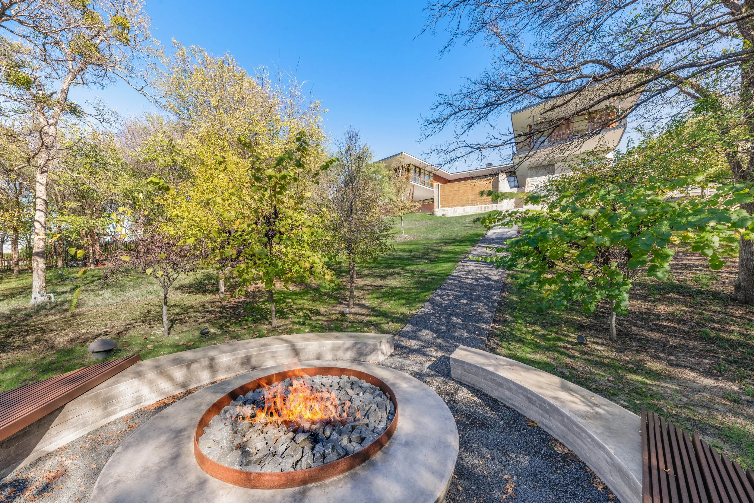 A modern building with large windows and balconies on a grassy hill, surrounded by trees with green and yellow leaves, with a stone pathway leading up to the house and a circular fire pit with burning fire in the foreground.