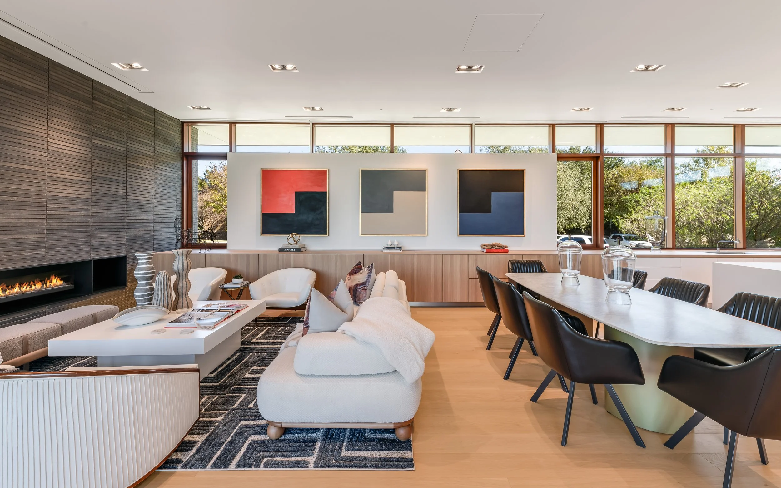 Modern living room and dining area with artwork, fireplace, and large windows showing greenery outside.