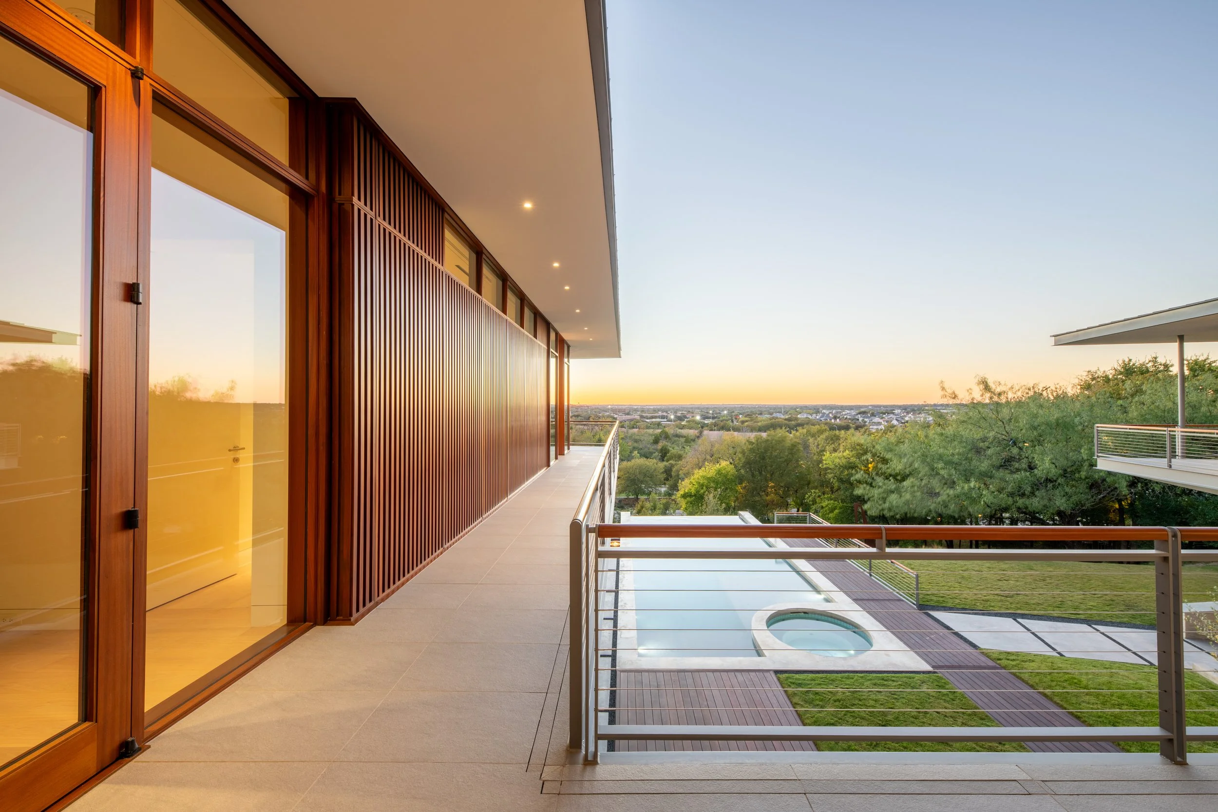 Modern balcony with glass doors, wooden panels, and a metal railing overlooking a landscaped yard with a hot tub and pool in the distance at sunset.