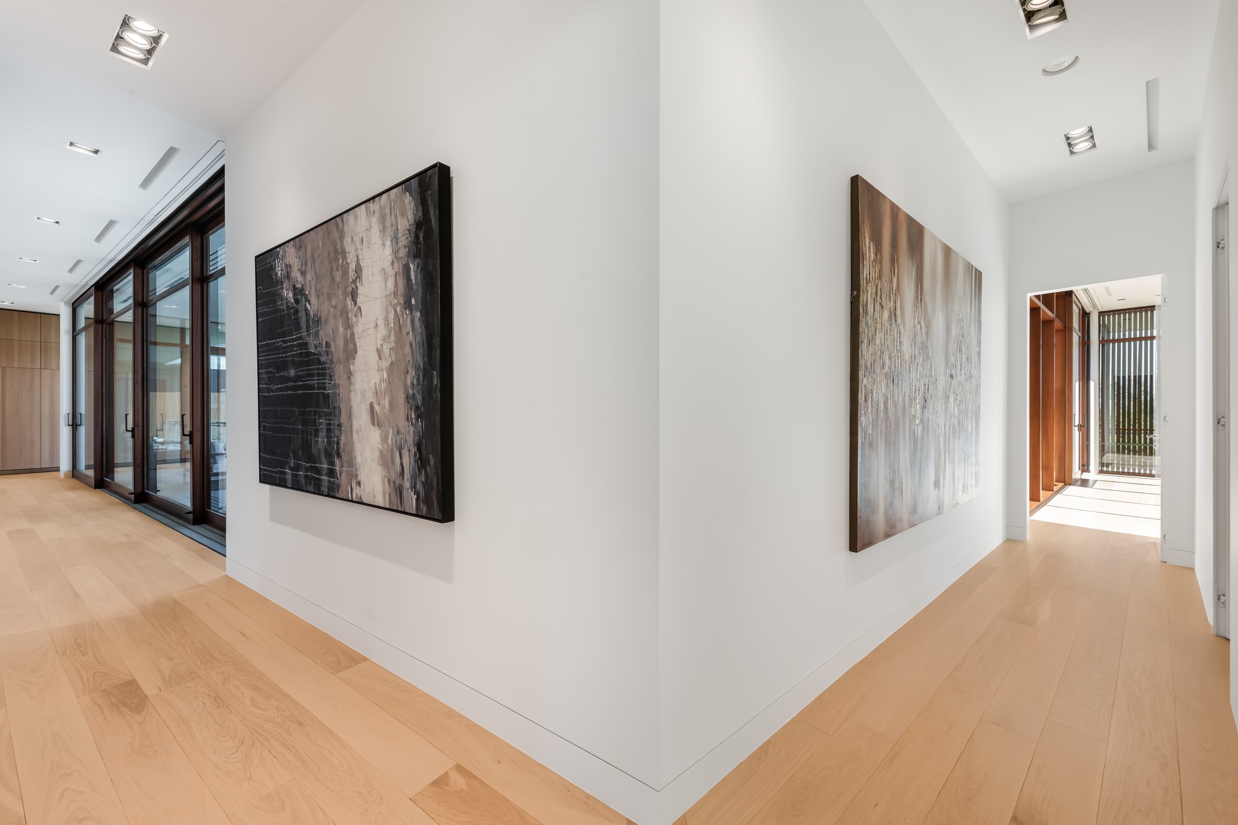 Modern art gallery with wooden floors, white walls, and abstract paintings on the walls. Large windows with black frames let in natural light.