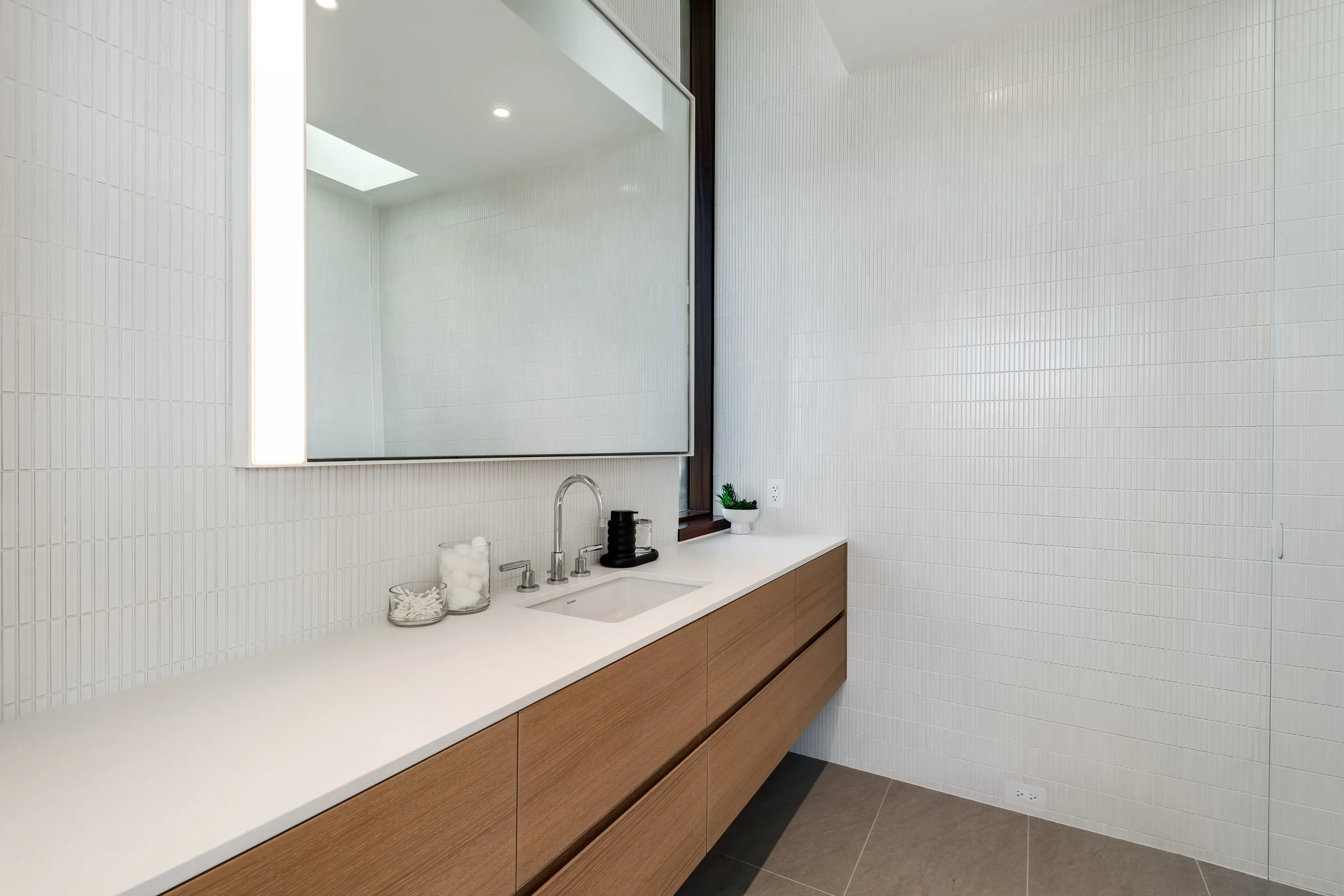 Modern bathroom with a white countertop, wooden cabinets, large mirror with backlighting, white tiled walls, and small decorative items near the sink.