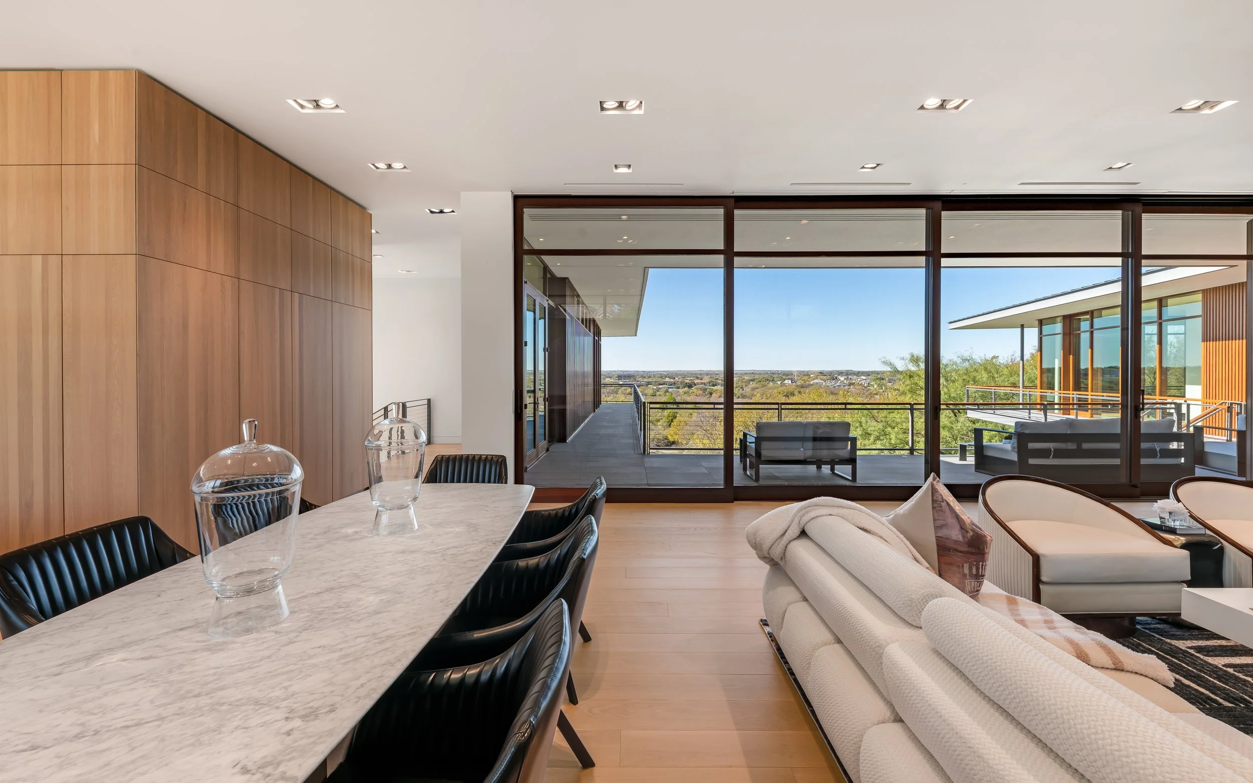 Modern living and dining room with wooden accents, large marble dining table, black chairs, beige sofa, and glass sliding doors leading to a balcony with outdoor seating and panoramic view.