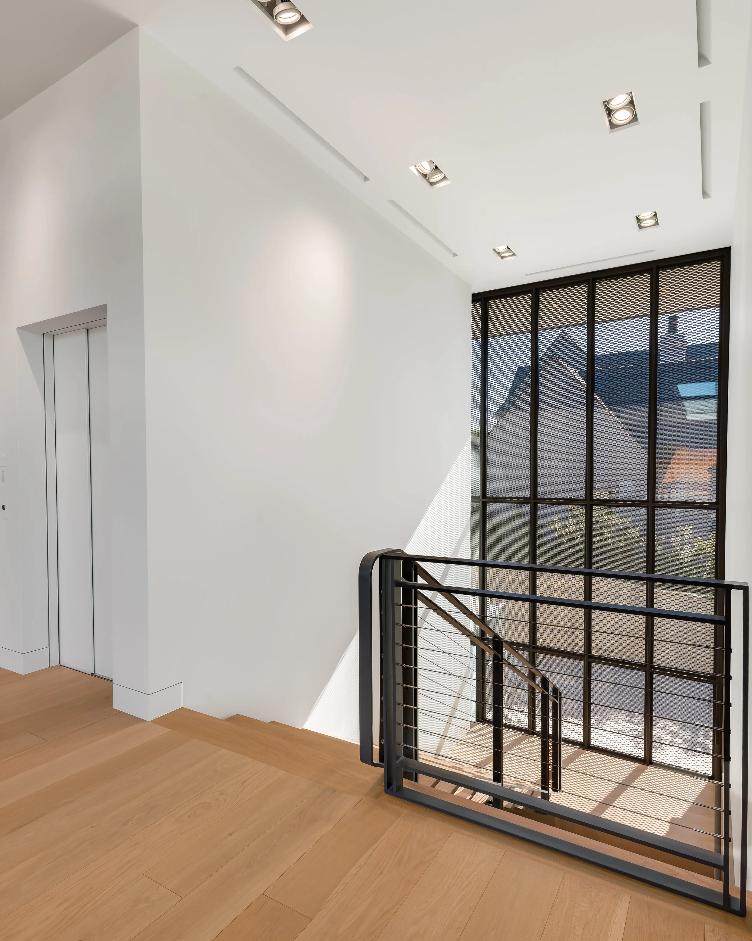 Modern interior staircase with black metal railing, large window with grid pattern showing neighboring houses, wooden floor, white walls, and ceiling with recessed lighting.