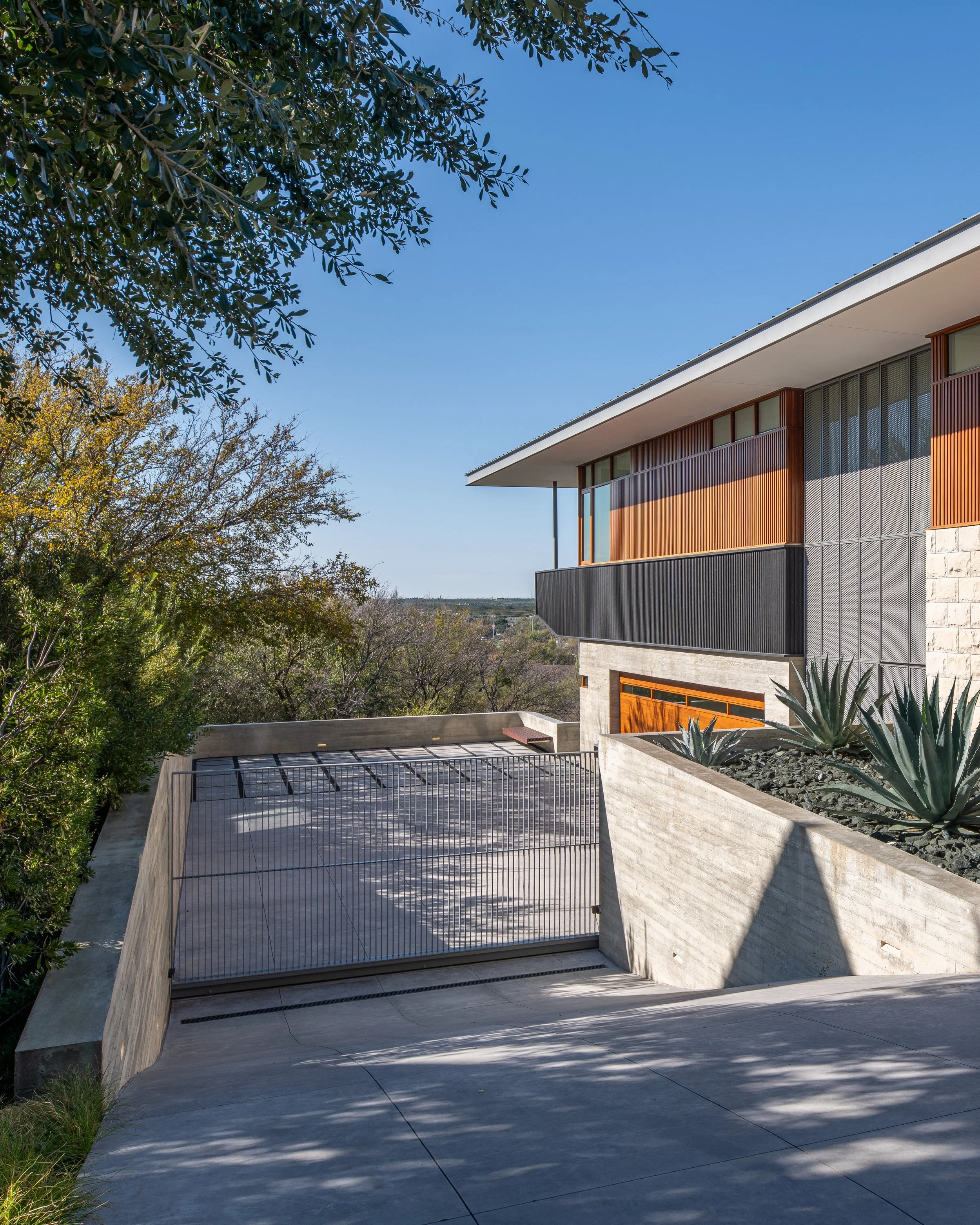 Modern house with wooden and concrete exterior, overlooking a driveway with a metal gate, surrounded by trees and desert plants under a clear blue sky.