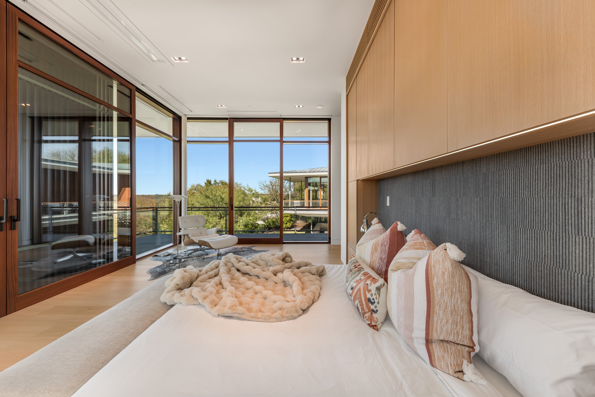 Modern bedroom with large windows, a bed with decorative pillows and a throw blanket, a balcony outside, and a black textured headboard.