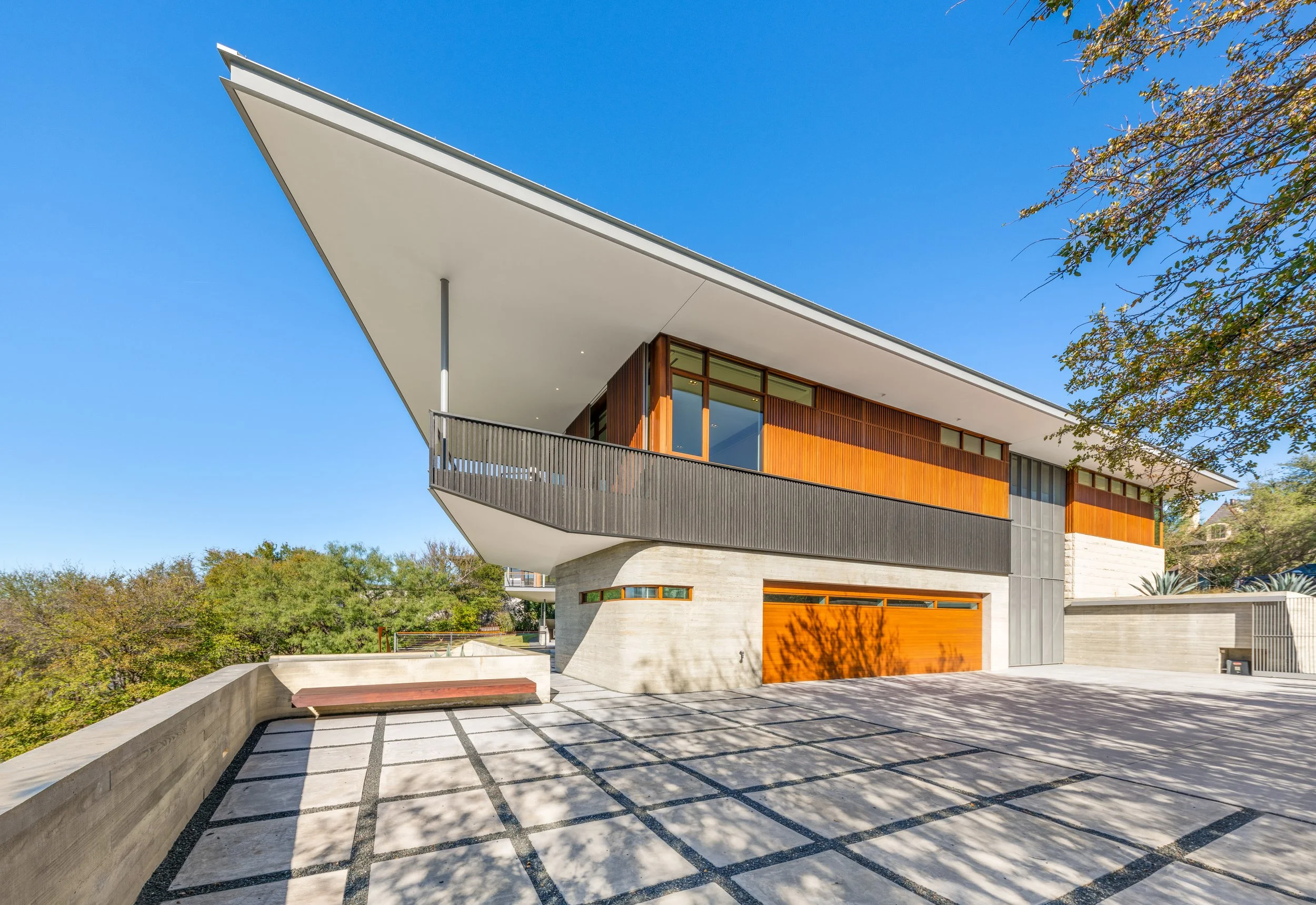 Modern house with a large overhanging roof, wooden paneling, large windows, and a concrete driveway with a bench and shadow patterns