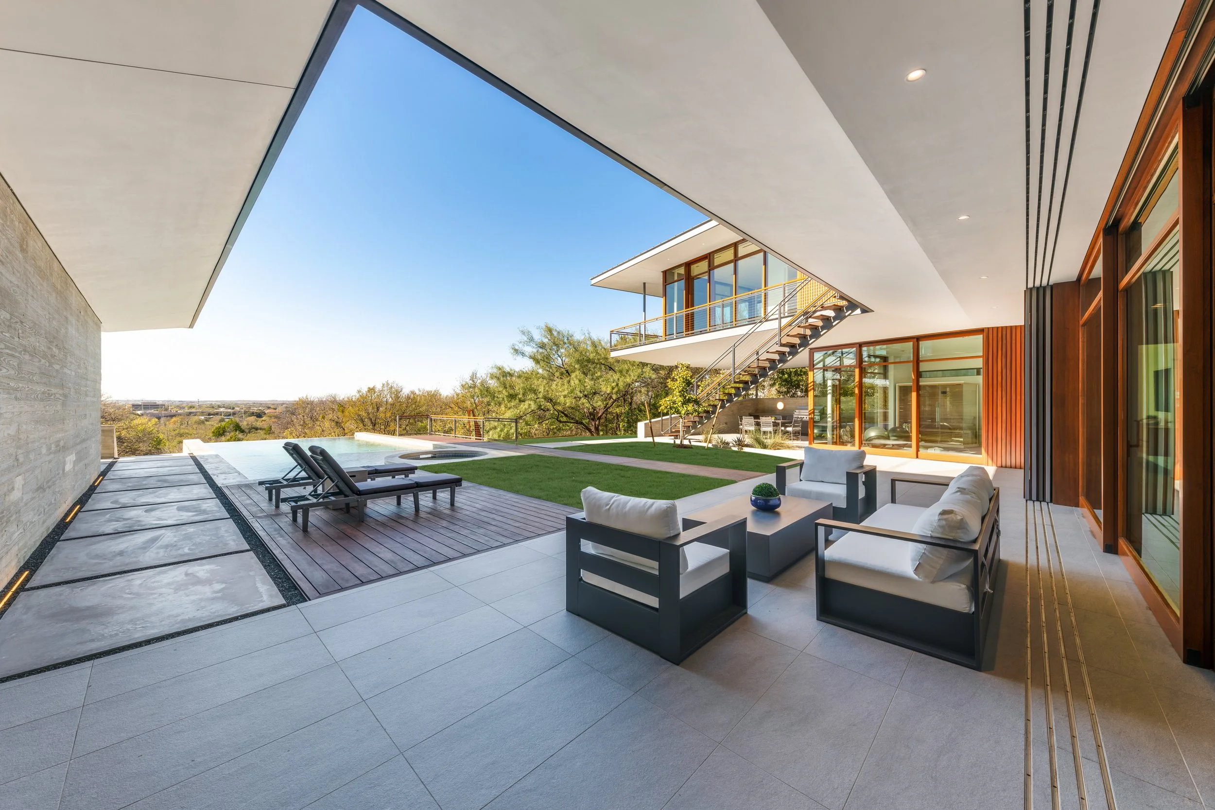Modern outdoor patio with seating area, lounge chairs, pool, and an open view of trees and sky.