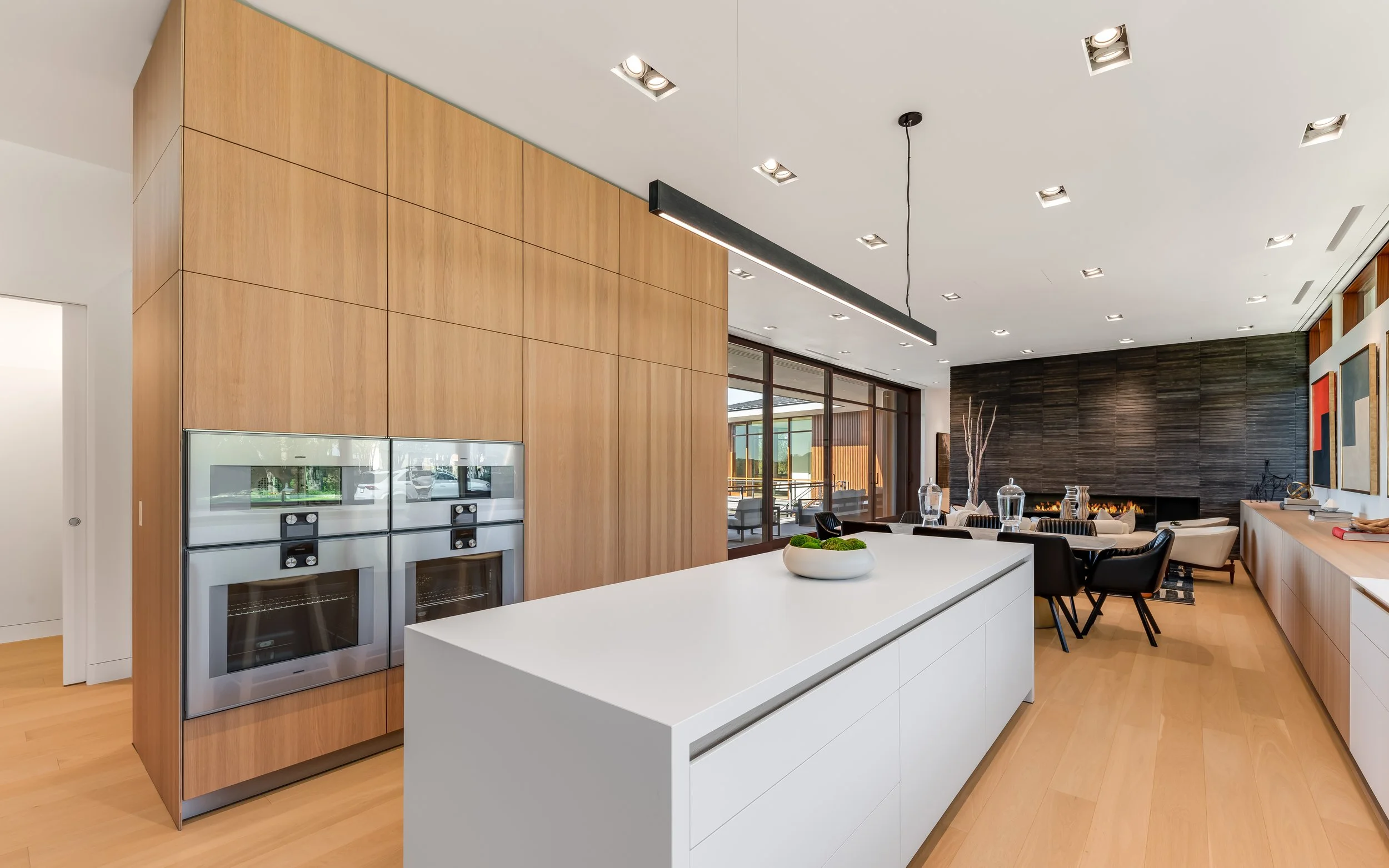 Open-concept modern kitchen and living room with wood and white finishes, black dining chairs, and a dark textured wall with a fireplace.