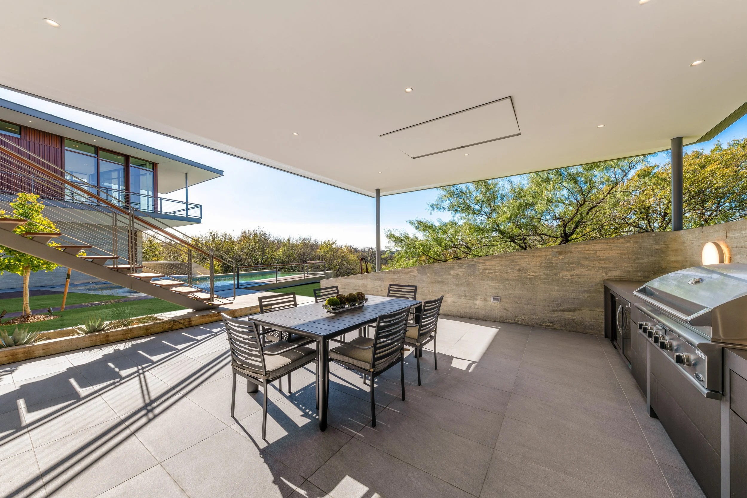 Outdoor patio with dining table and chairs, built-in grill, and a view of trees and neighboring house.
