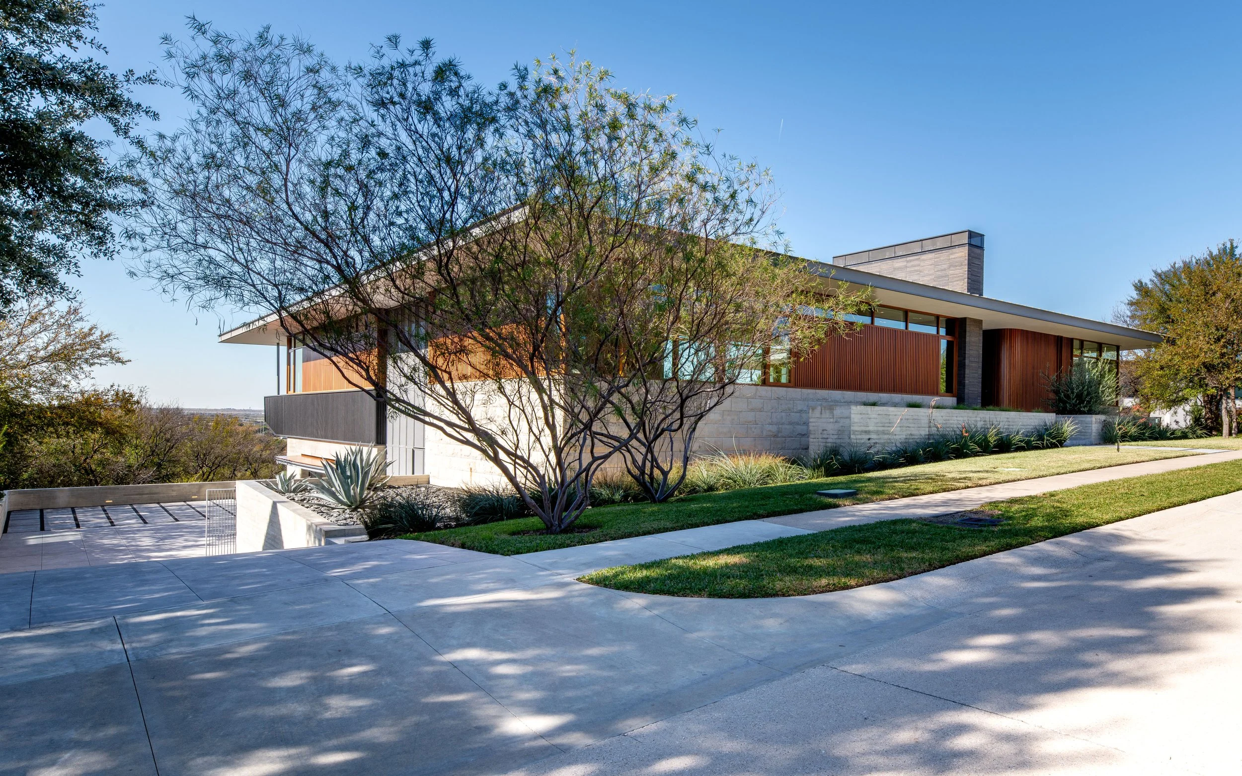 Modern house with large windows, stone and wood exterior, surrounded by trees and a neatly landscaped yard under a clear blue sky.