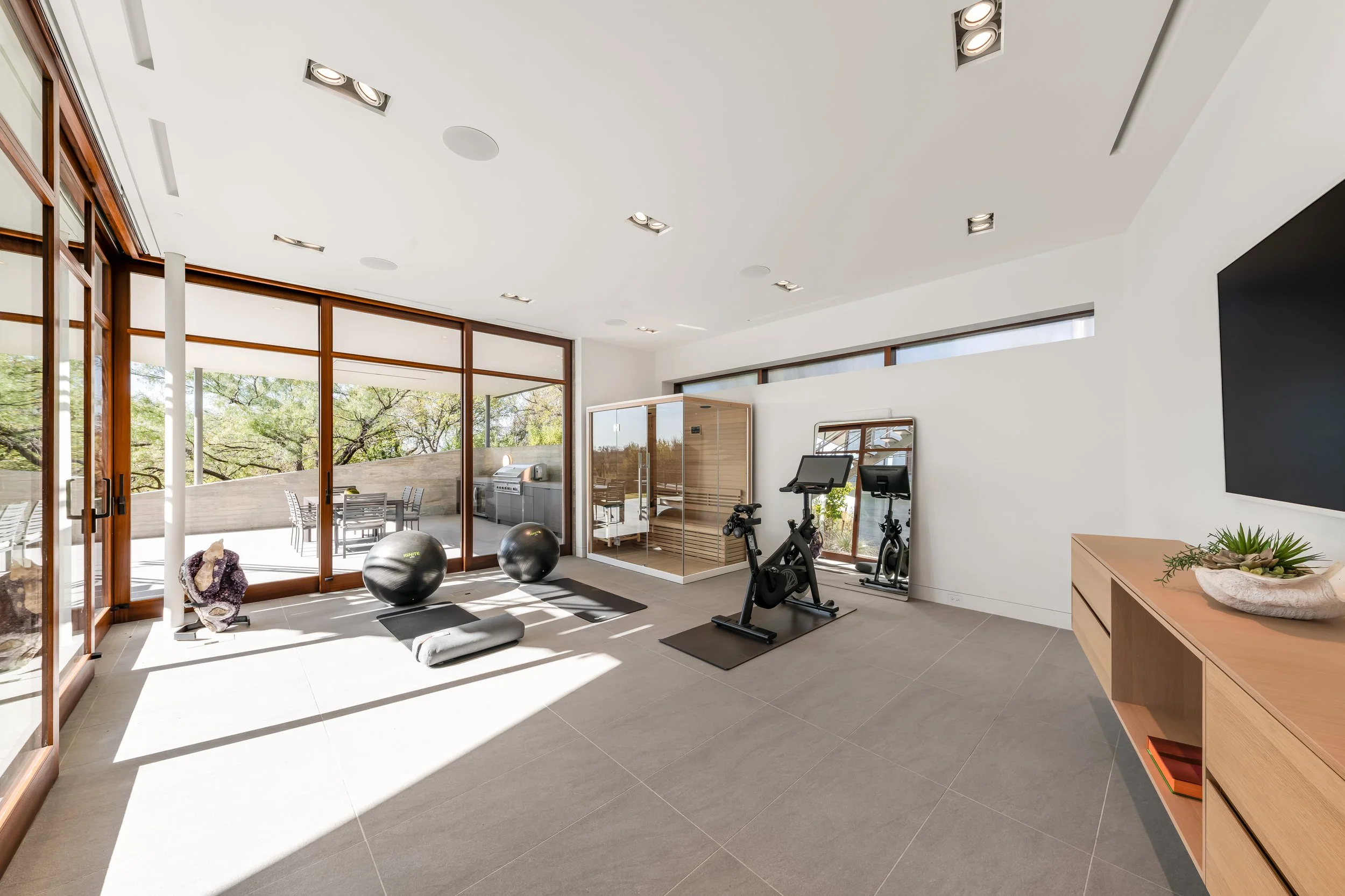 Modern home gym with exercise bike, yoga mats, exercise balls, and infrared sauna, with large windows showing trees outside.