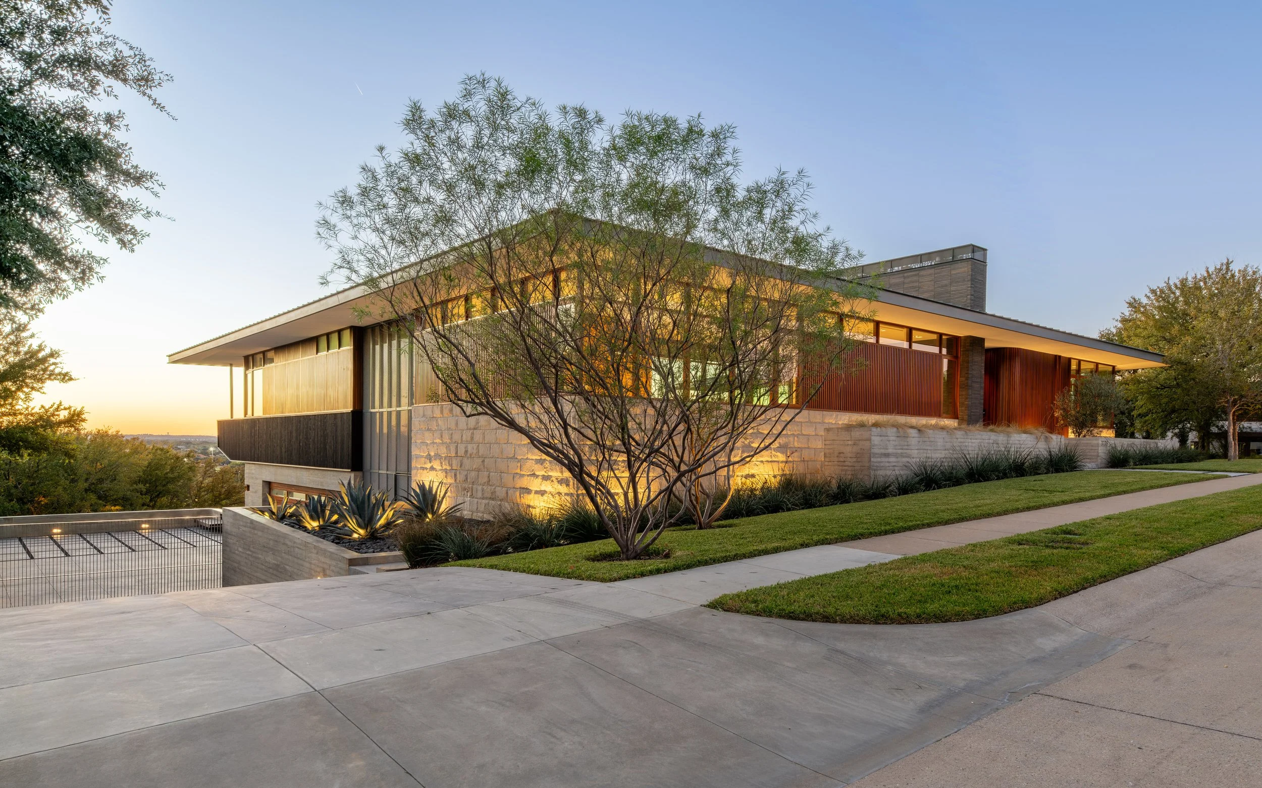 Modern house with large windows, a sloped roof, and exterior lighting, surrounded by trees and a landscaped yard at dusk.