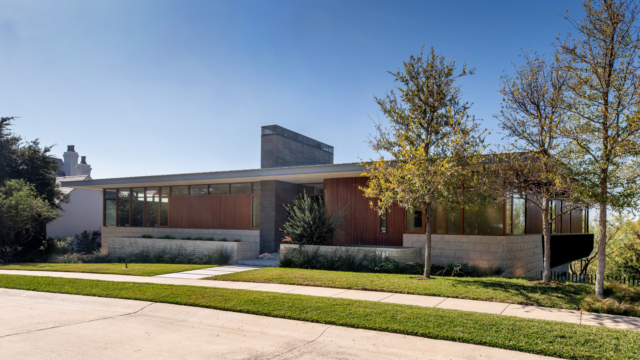 Modern house with large windows, wooden and stone exterior, surrounded by trees and a well-maintained grassy lawn, on a sunny day.