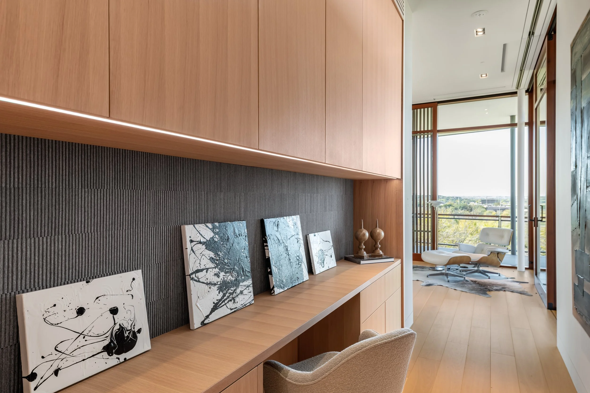 Modern interior with wooden cabinets, abstract artwork, and a view of a balcony with a lounge chair and large window.