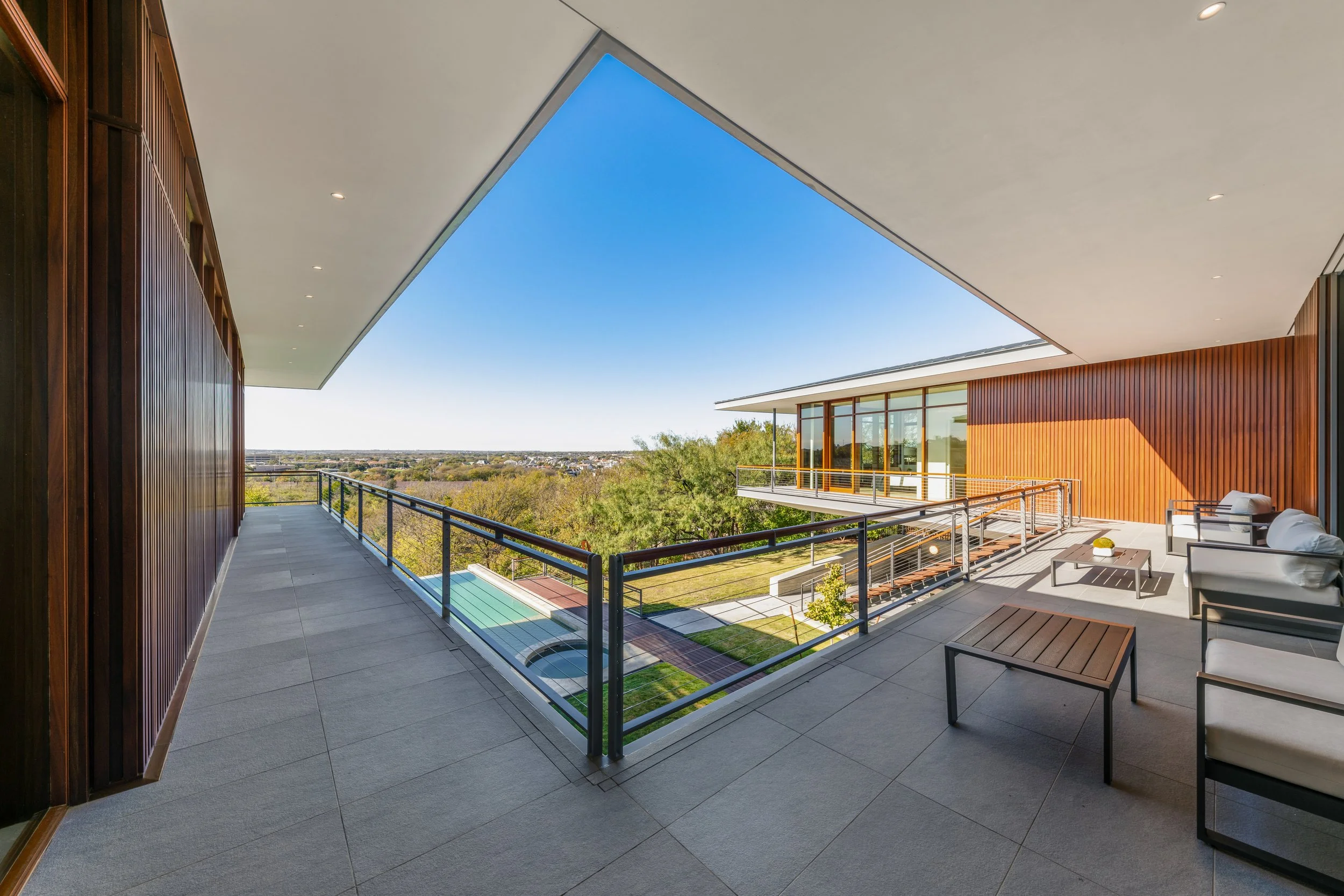 Large modern balcony with outdoor seating and glass railing, overlooking a suburban landscape with trees and blue sky.